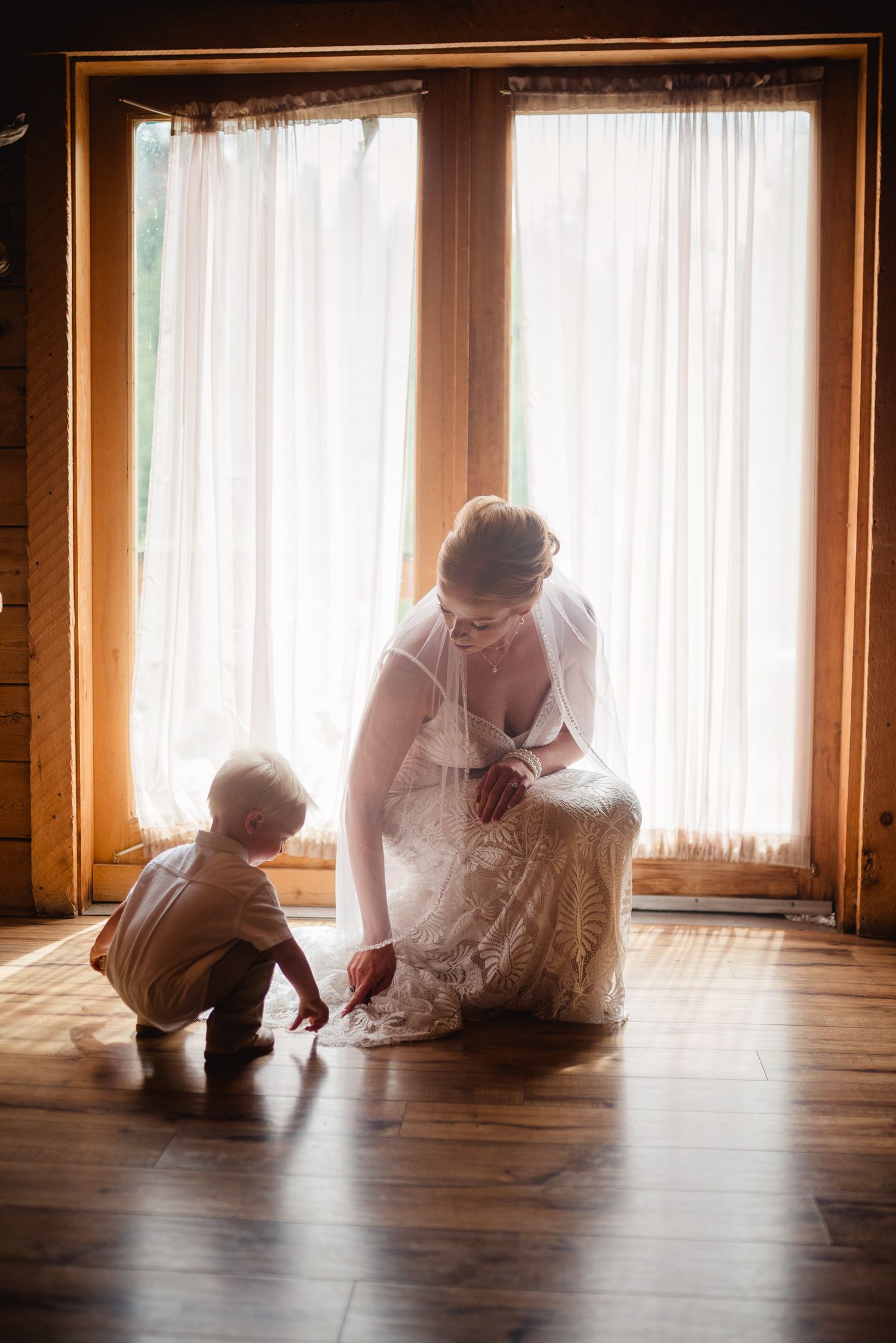 A bride in a white lace wedding dress and veil kneels with a young toddler boy on a warm wooden floor by large windows.