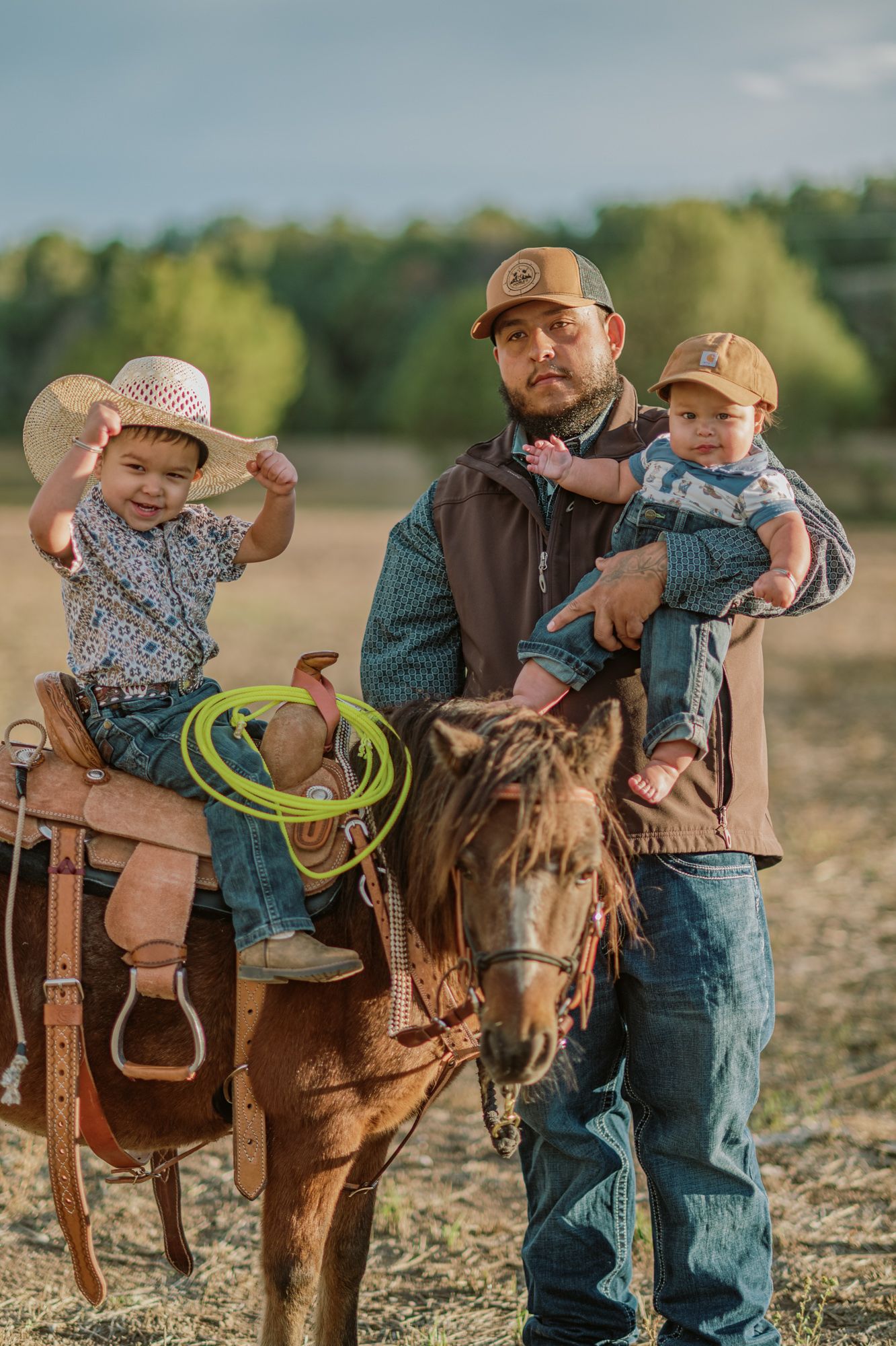 A father in a cap holds a baby while a young child in a cowboy hat sits on a saddled horse in a rural field.