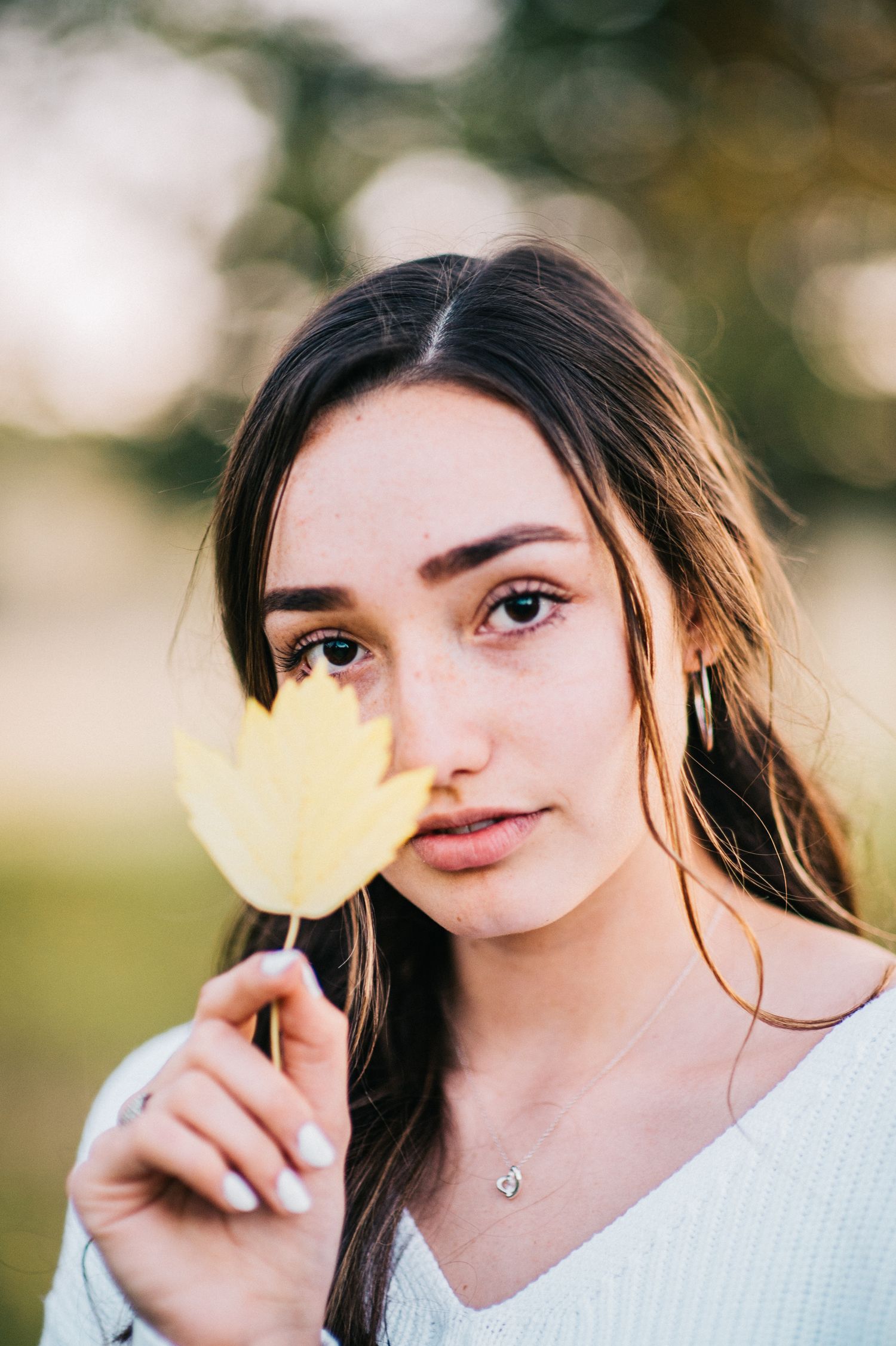 Person holding yellow leaf in front of face while wearing white sweater in natural setting.