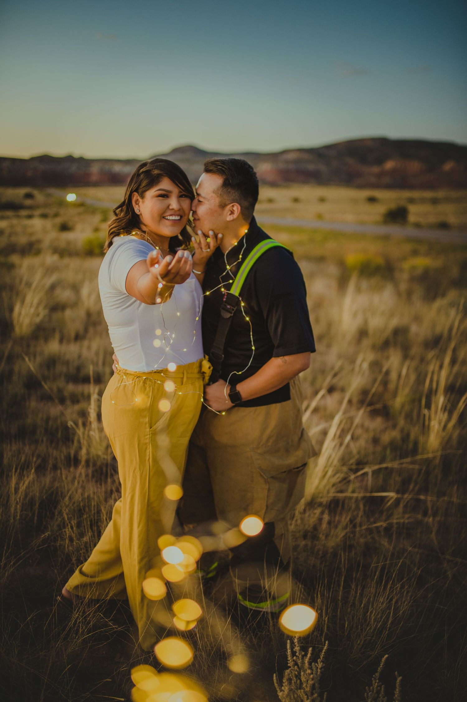 Playful couple laughing in desert field with bokeh light effects at sunset.