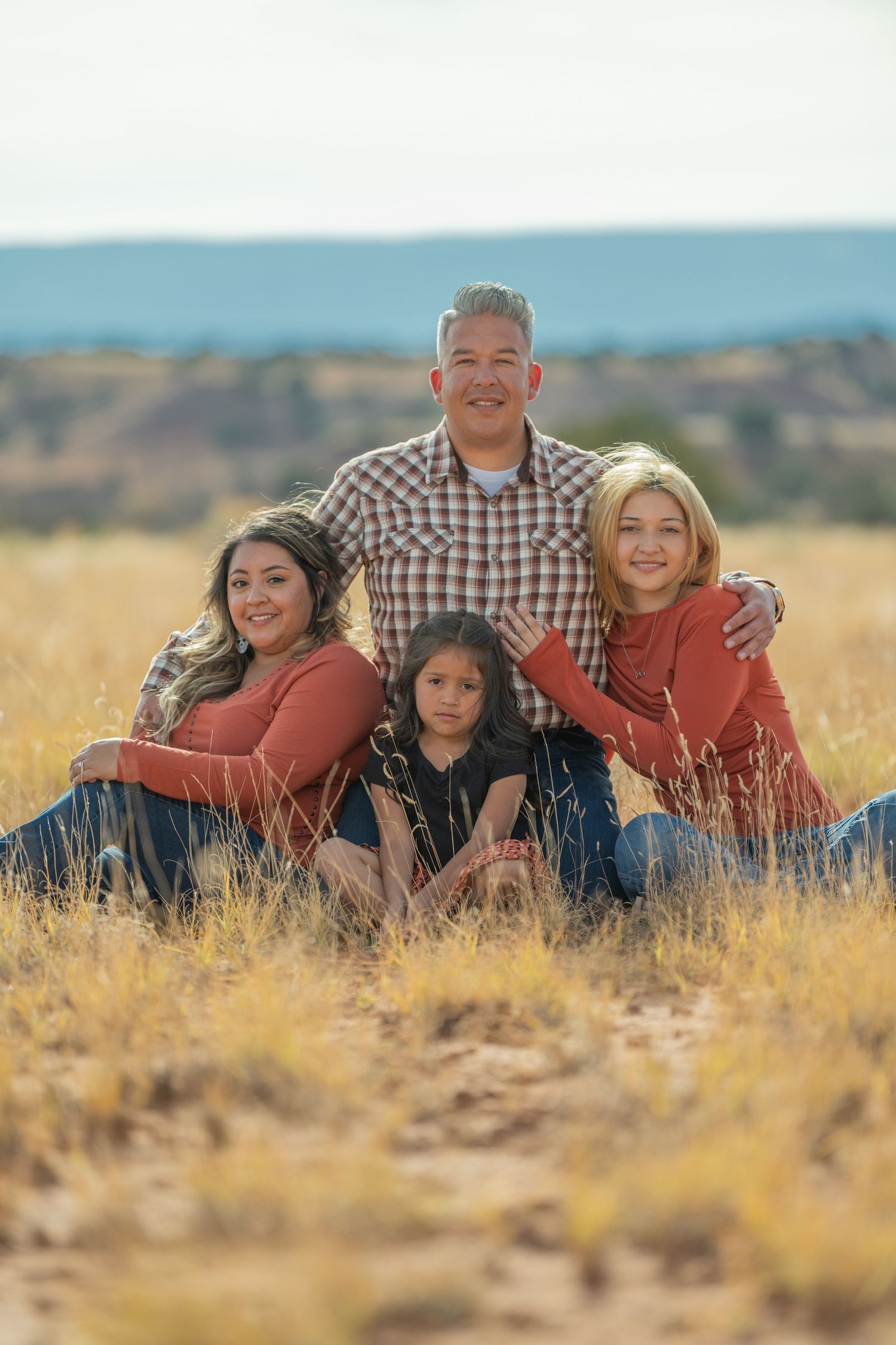 A family sits together in a golden field during sunset wearing coordinated red and plaid outfits against a desert landscape.