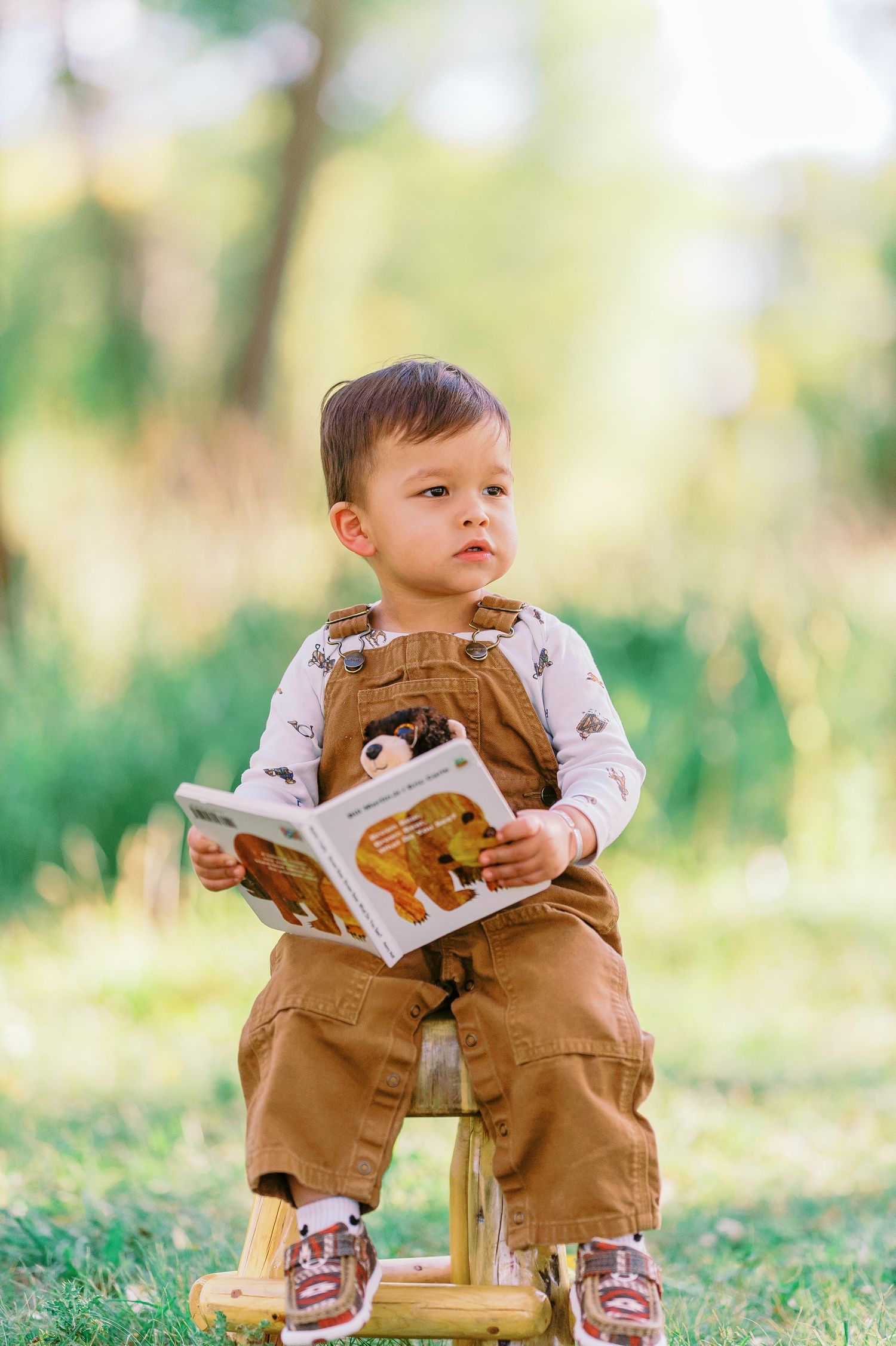 Small child in brown overalls sits on wooden stool outdoors while holding a picture book in a natural setting.