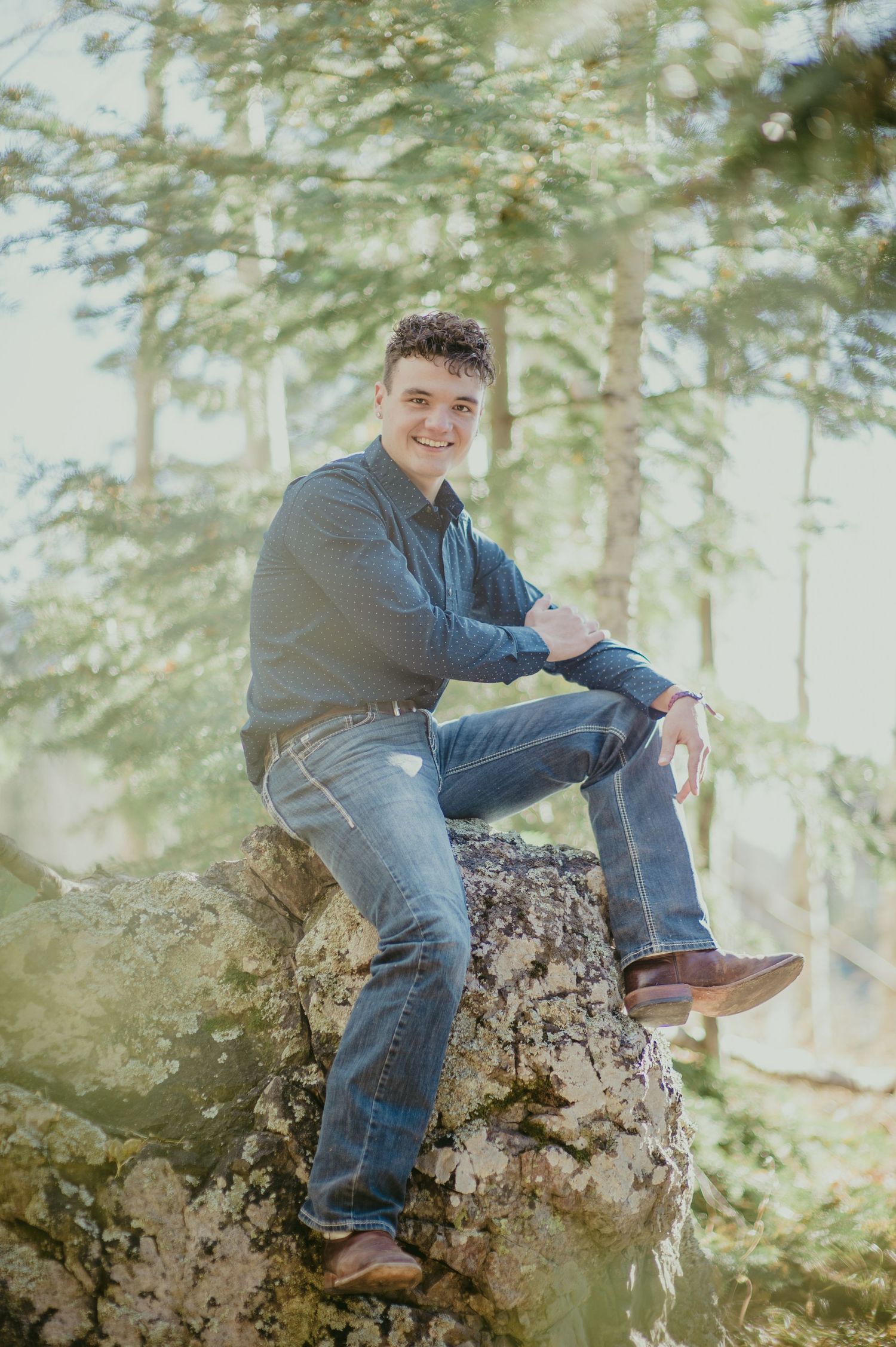 Young man in blue shirt and jeans sitting on a mossy rock in a sunlit forest, smiling for senior photos.