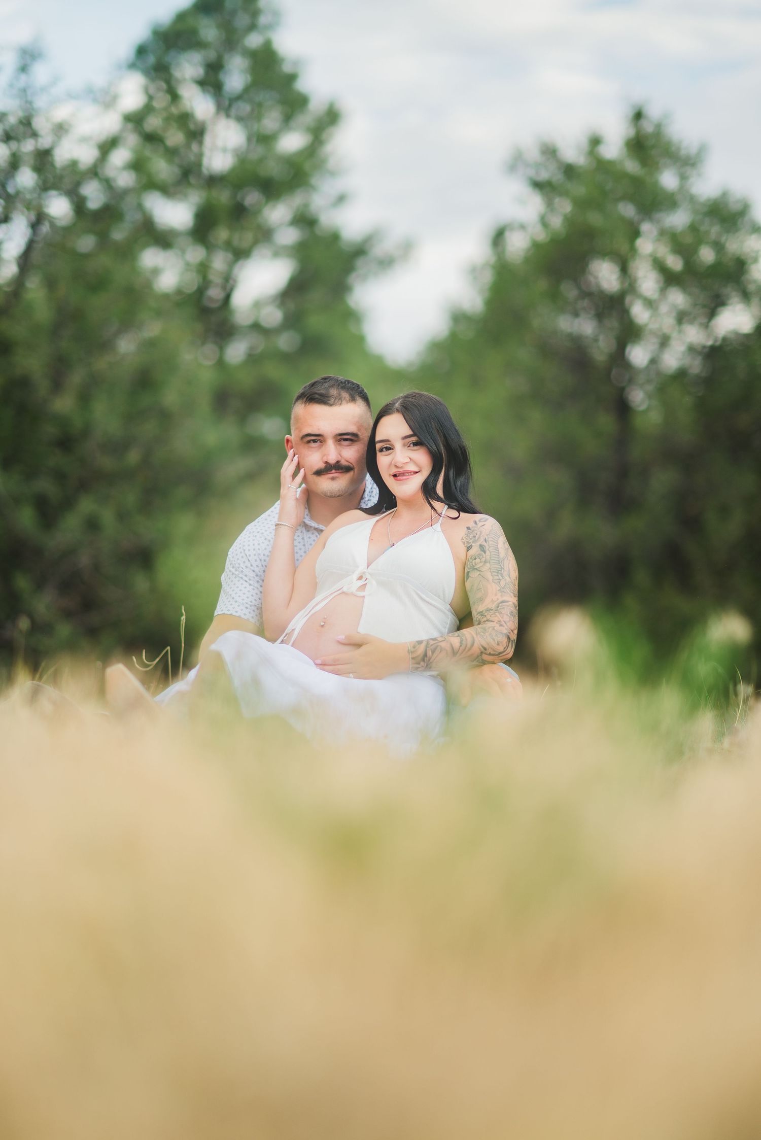 Couple sitting in golden grass field, pregnant woman with tattoos in white dress, man with mustache, green trees background.