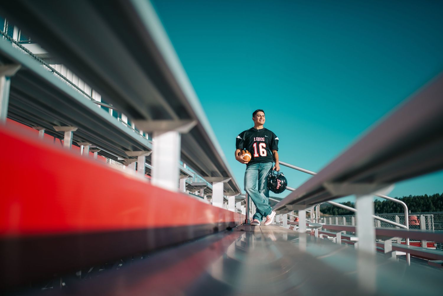 Football player #16 in black jersey stands on stadium bleachers under a bright blue sky.