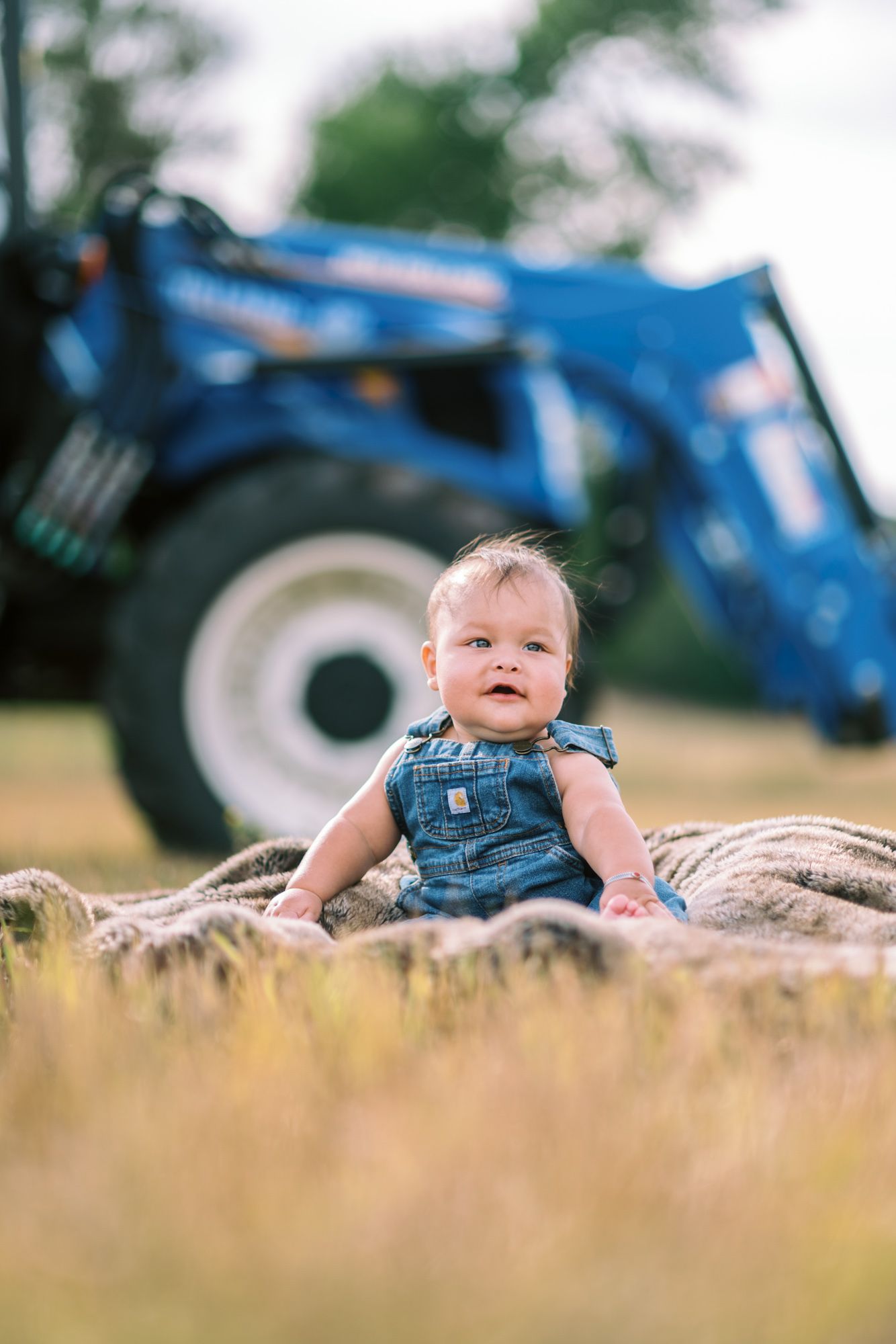 Adorable baby in denim overalls sitting on a hay bale in a field with a blue tractor in the background.