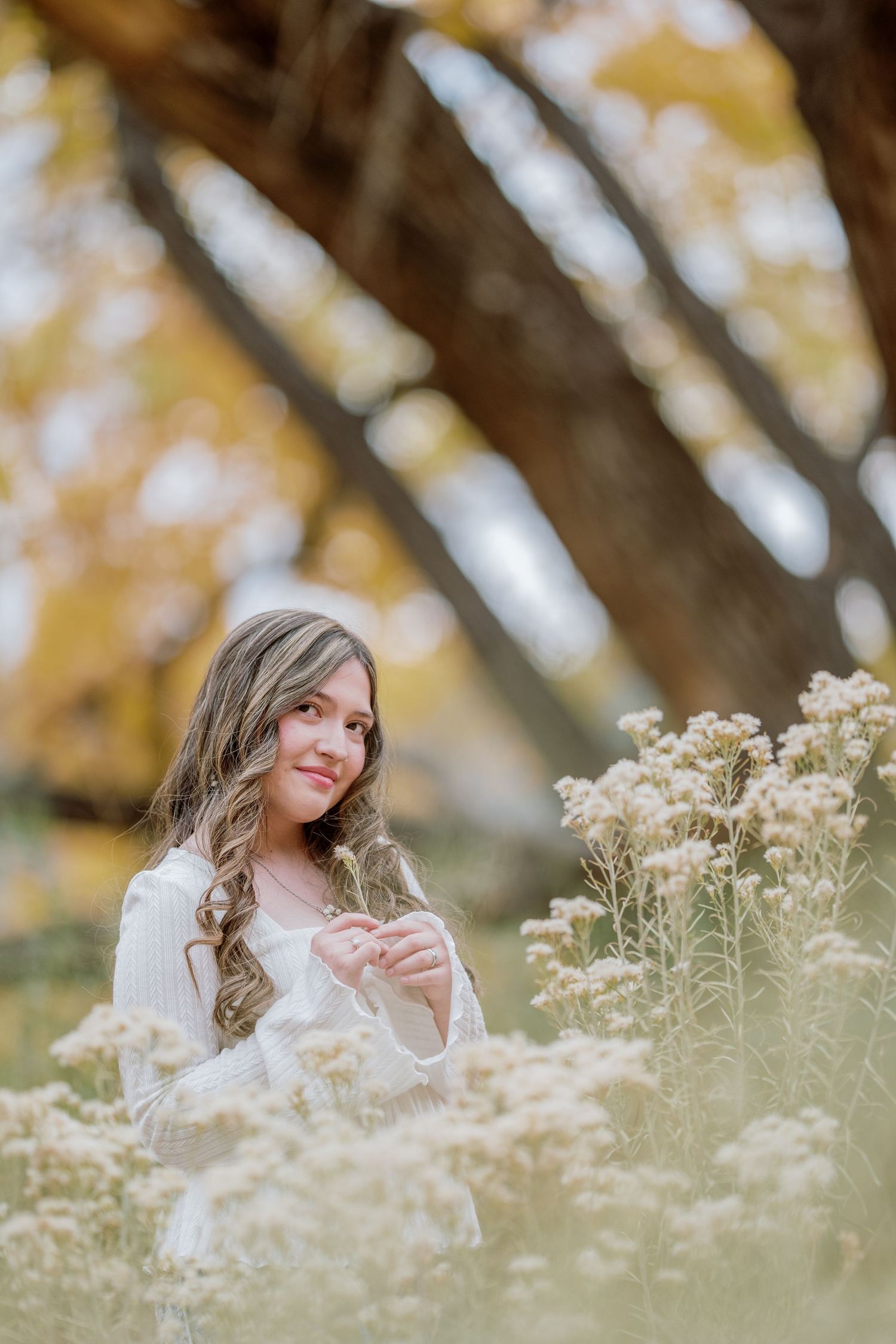 A woman in white sits among delicate wildflowers on an autumn day with golden bokeh in the background.