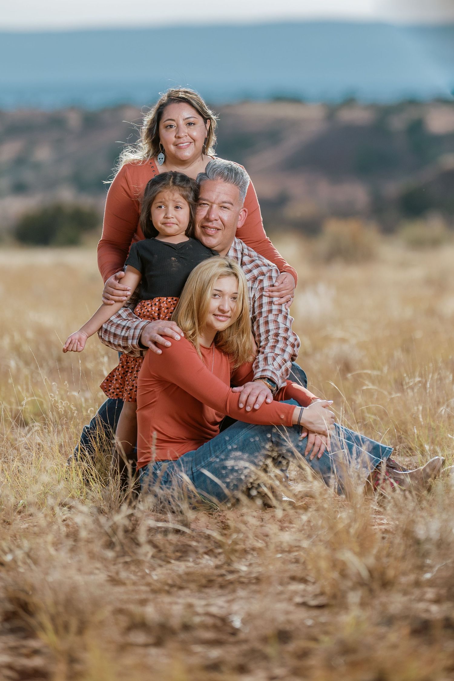 A family poses together in a golden field with mountains visible in the distant background during sunset.