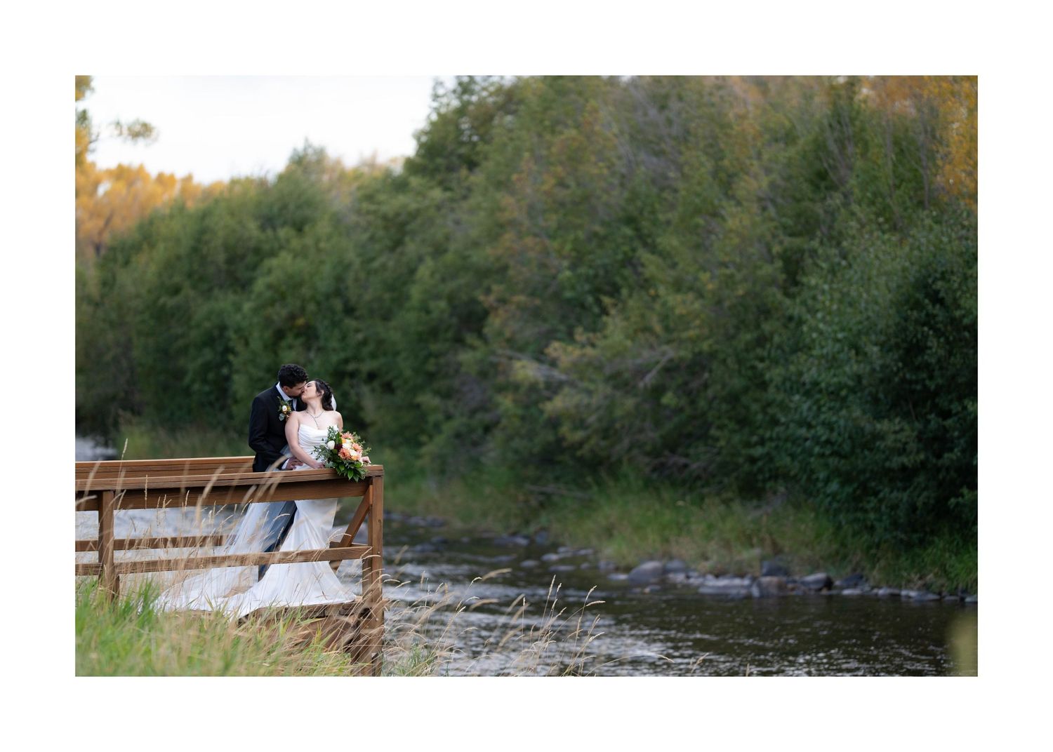 A couple sits together on a wooden bridge overlooking a tranquil lake surrounded by lush green forest.