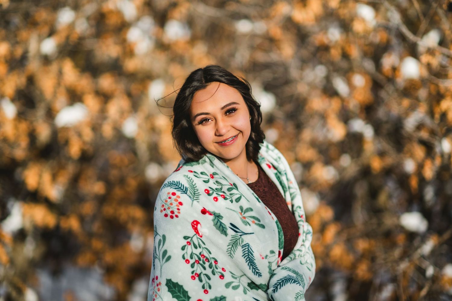 A person in a white floral dress poses outdoors with autumn foliage in the background.