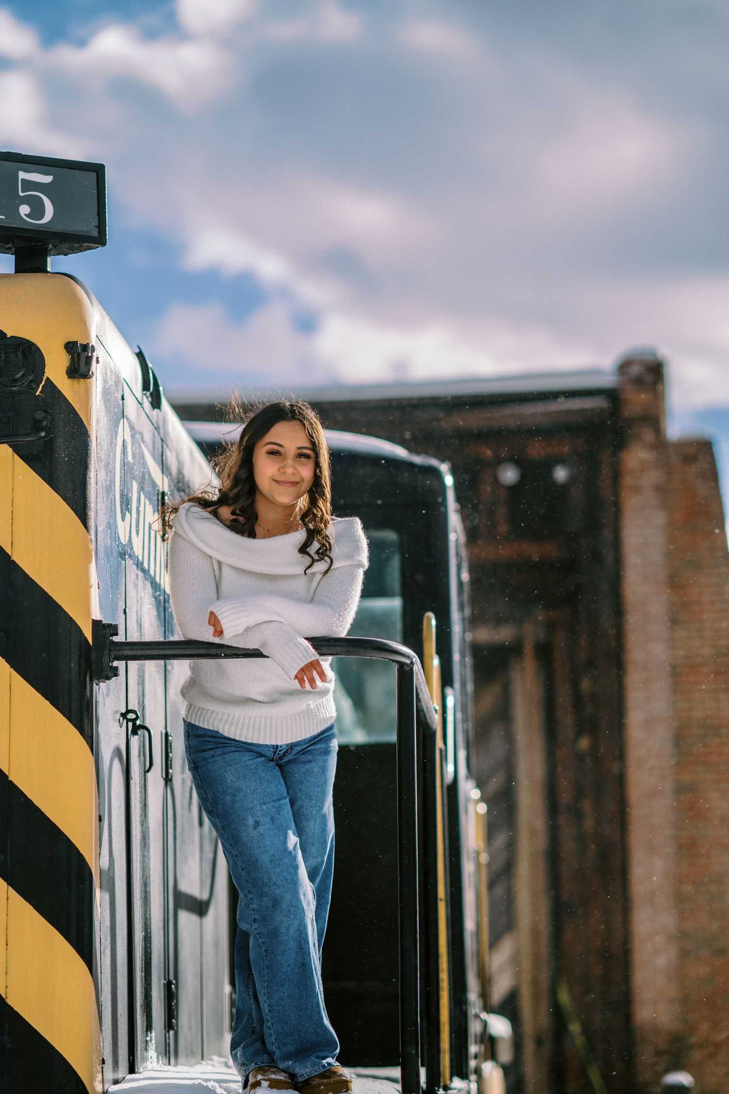A person in a white sweater and jeans poses on a train platform with brick buildings in the background.