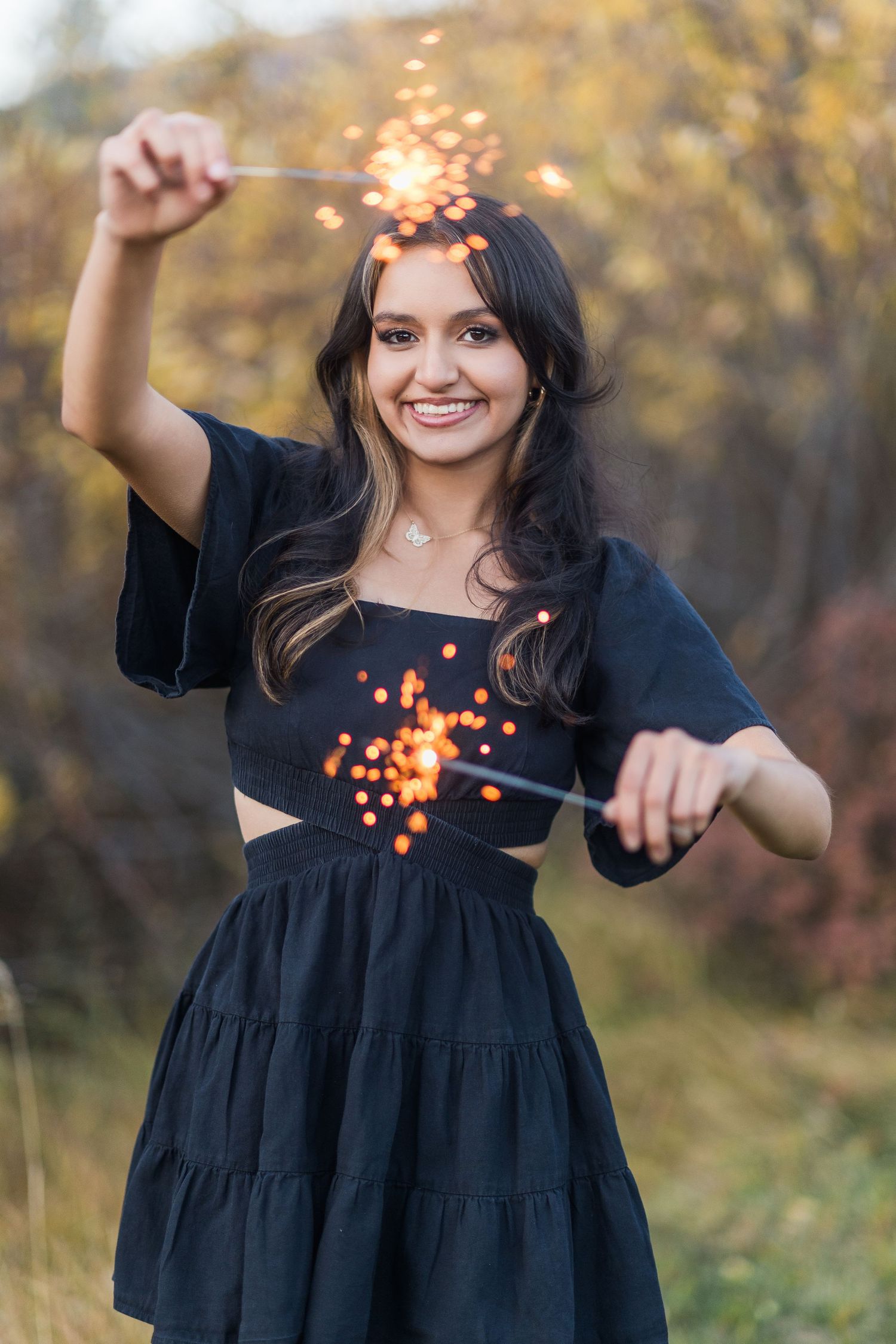 A woman in a flowing navy dress tosses orange glitter in an autumn outdoor setting, creating a magical effect.