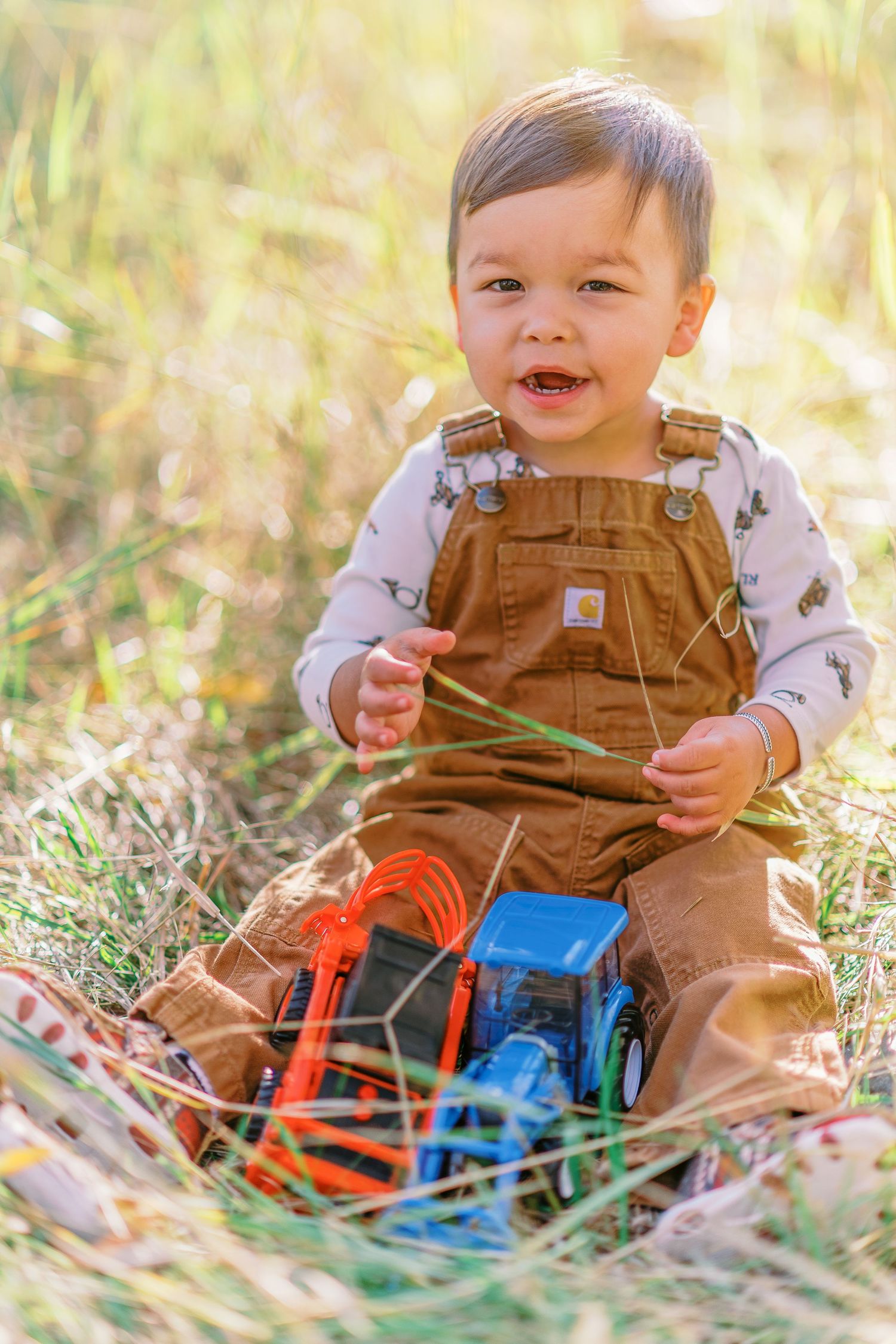 Baby in brown overalls plays with colorful toys while sitting in sunlit grass on an autumn day.
