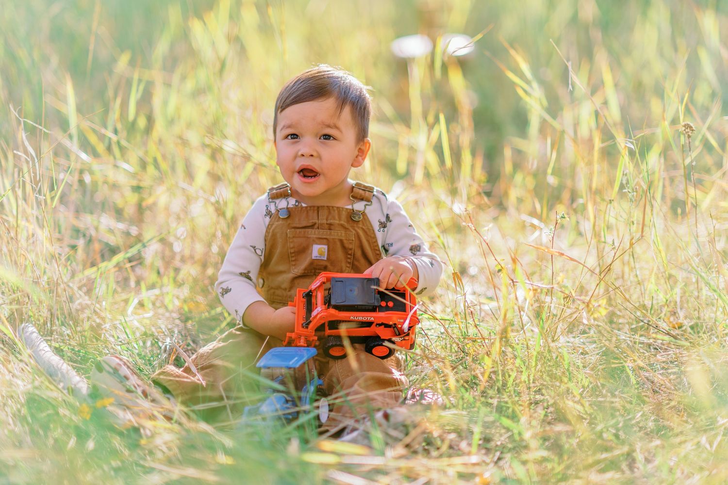 A child plays in a golden autumn field with a toy camera while wearing a brown vest and orange shirt.