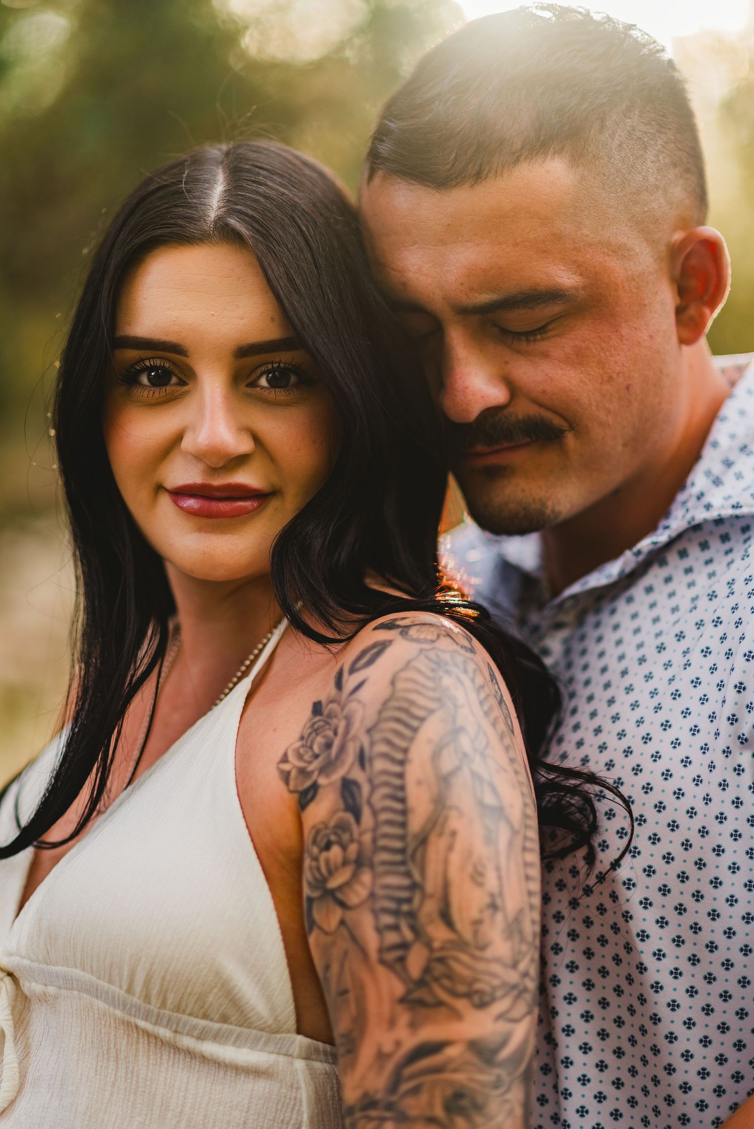 Couple posing outdoors in golden light, woman with tattoo sleeve in white dress, man in patterned shirt, romantic portrait.