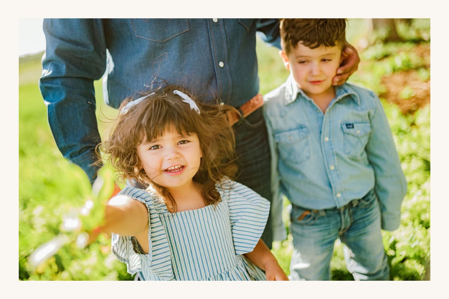 Two young children smiling outdoors in a green field, girl in striped dress and boy in denim shirt.