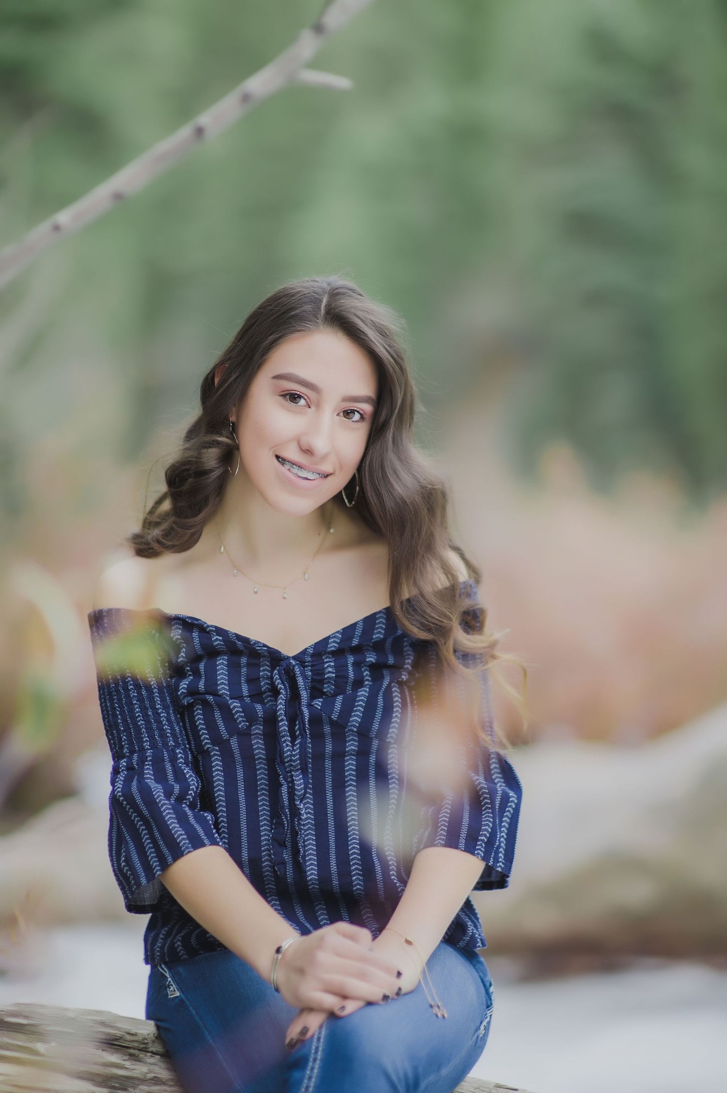 Smiling young woman in blue off-shoulder top sitting on log in natural outdoor setting.