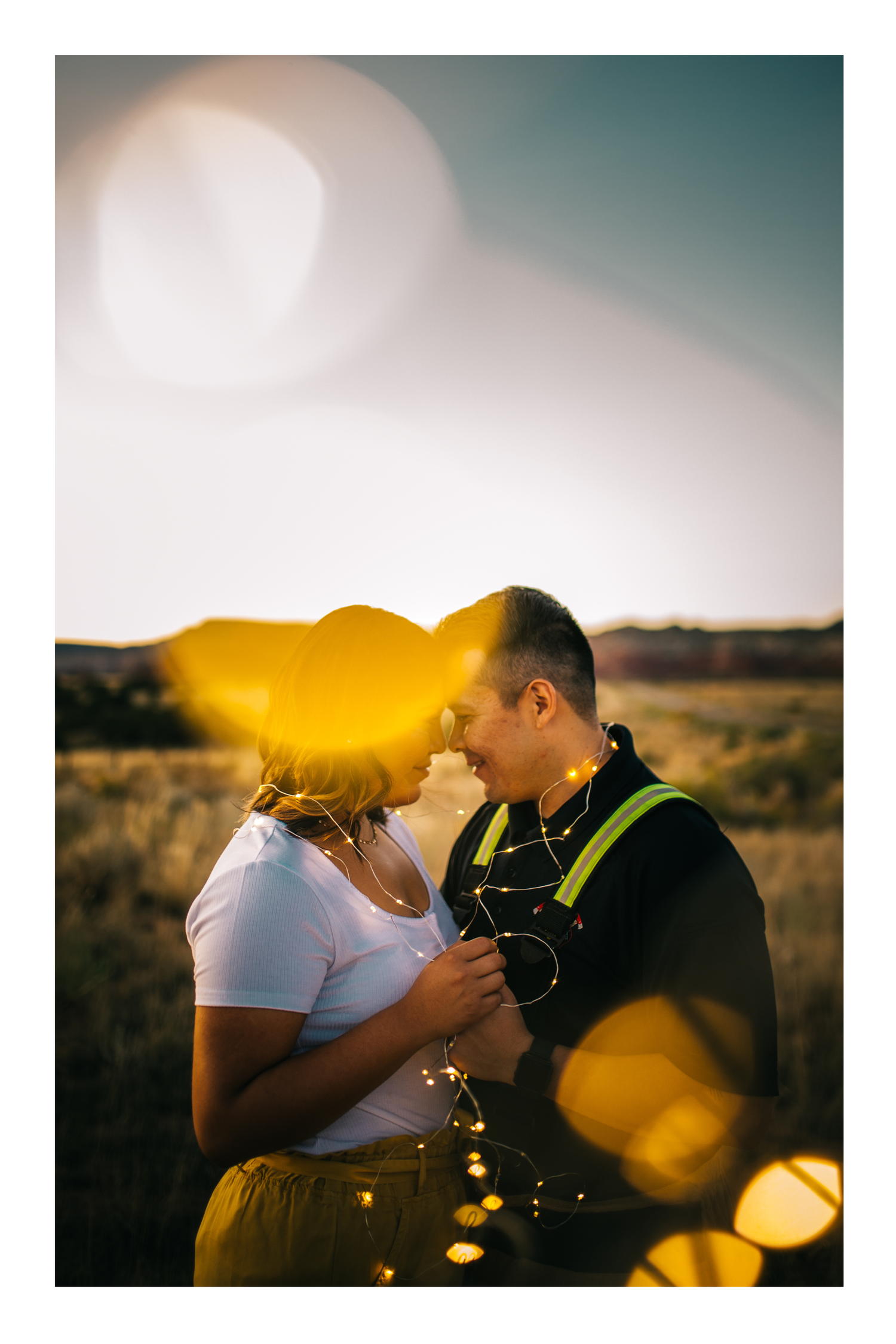 Couple sharing a romantic moment in a golden field at sunset, wrapped in fairy lights with warm lens flare effects.