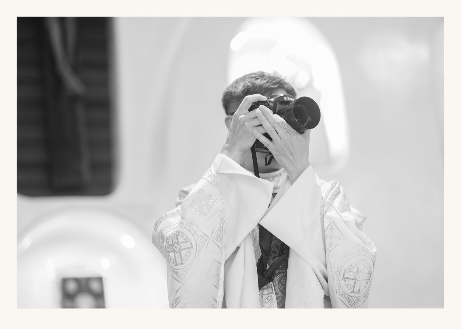 A priest in white vestments holds a professional camera up to his face during a church ceremony.