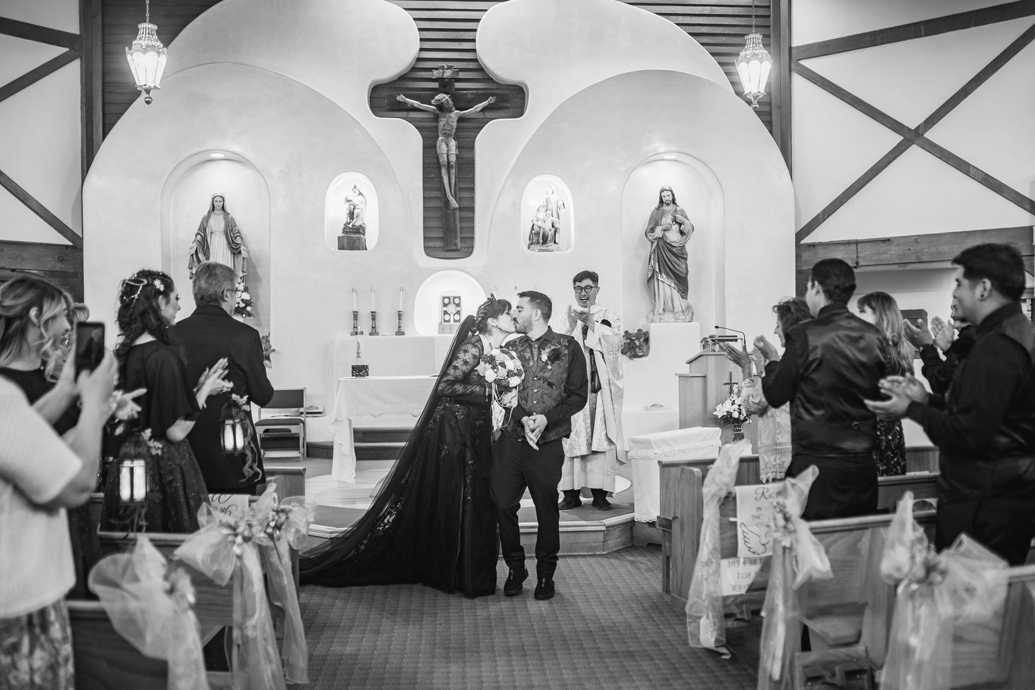Black and white photo of a couple kissing at the altar during a Catholic church wedding ceremony with guests watching.