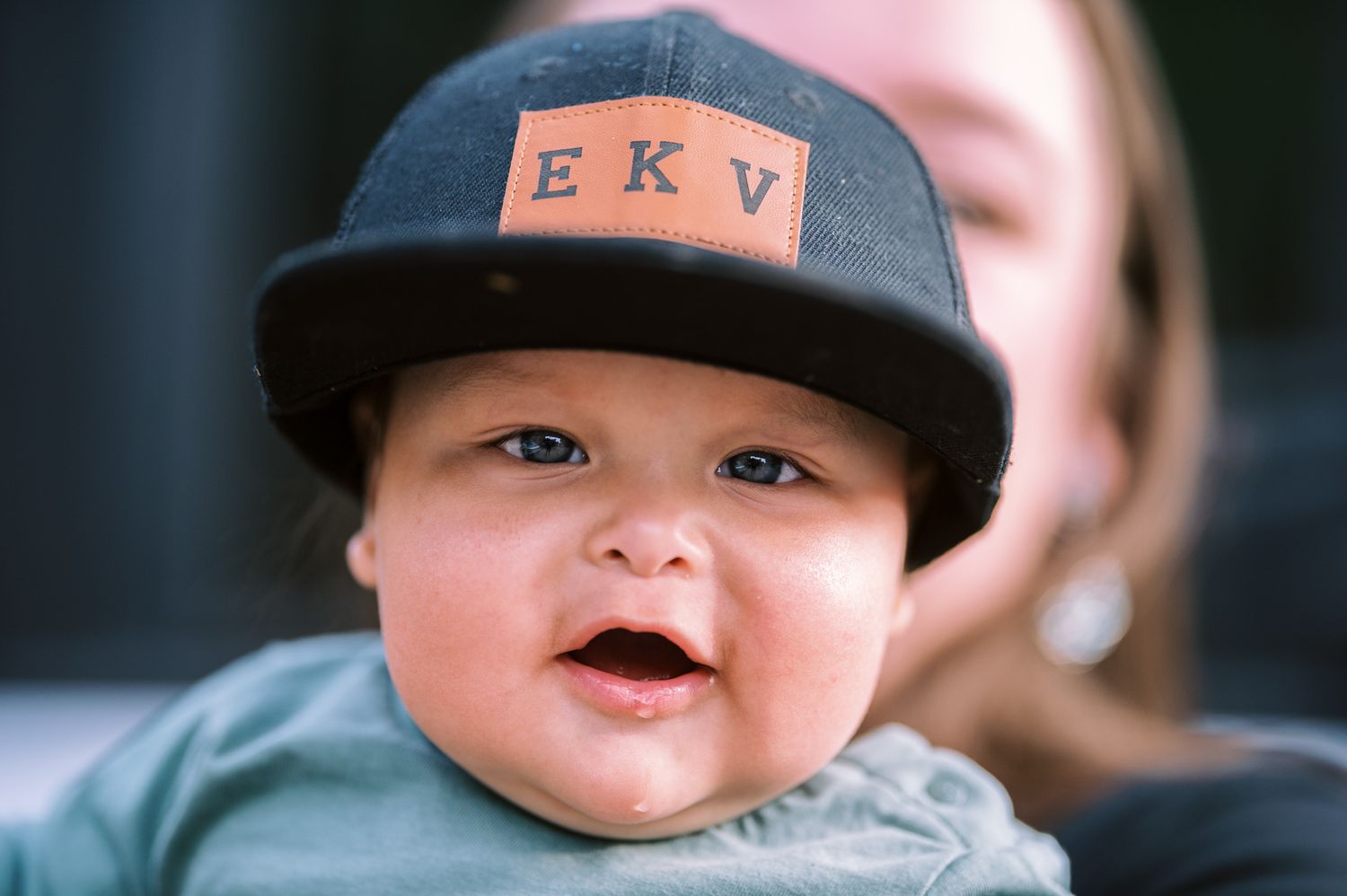 Adorable baby wearing a black snapback hat with EKV monogram patch, smiling with mouth open.