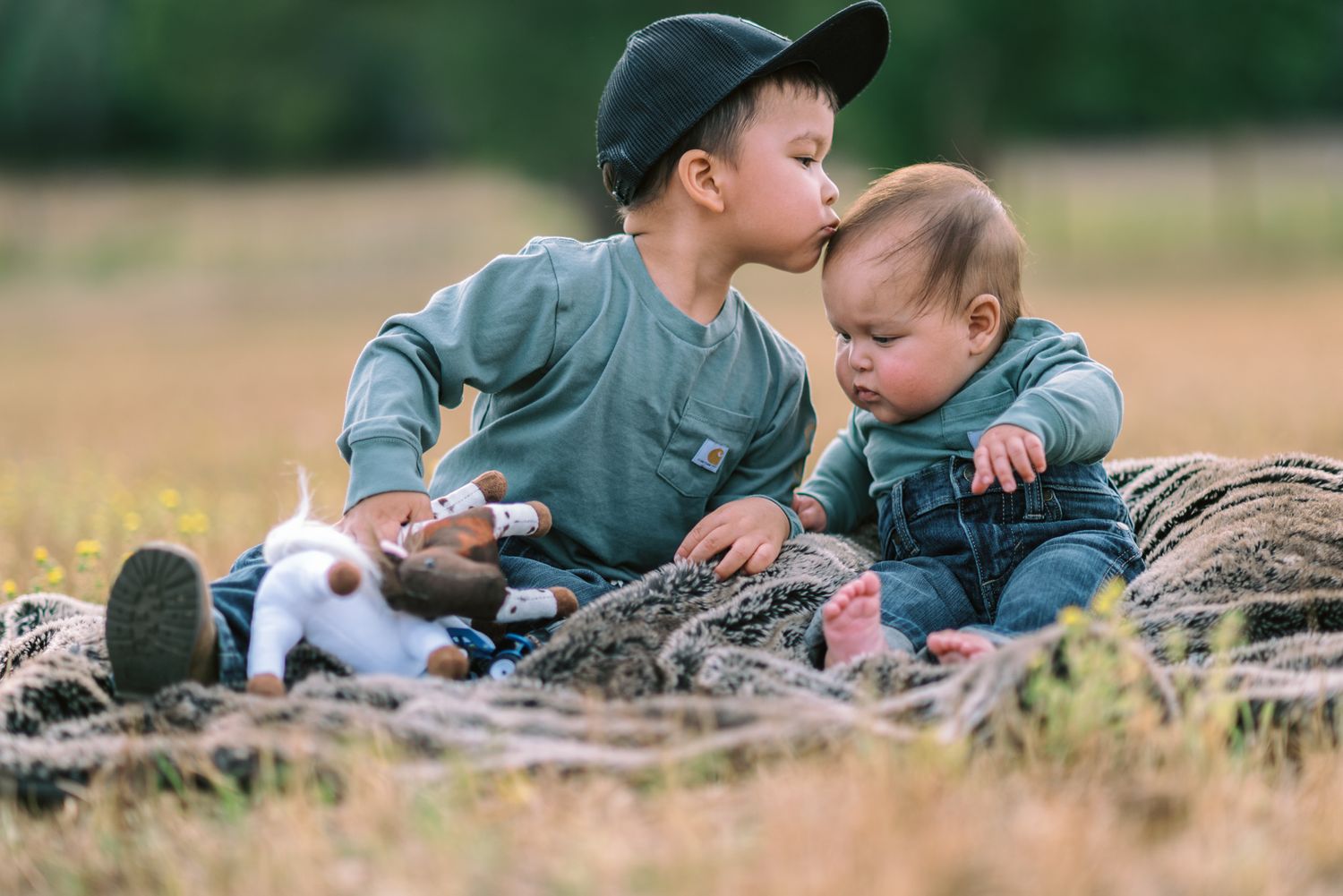 Two young children in matching green outfits sit on a blanket outdoors with a small puppy on a sunny day.