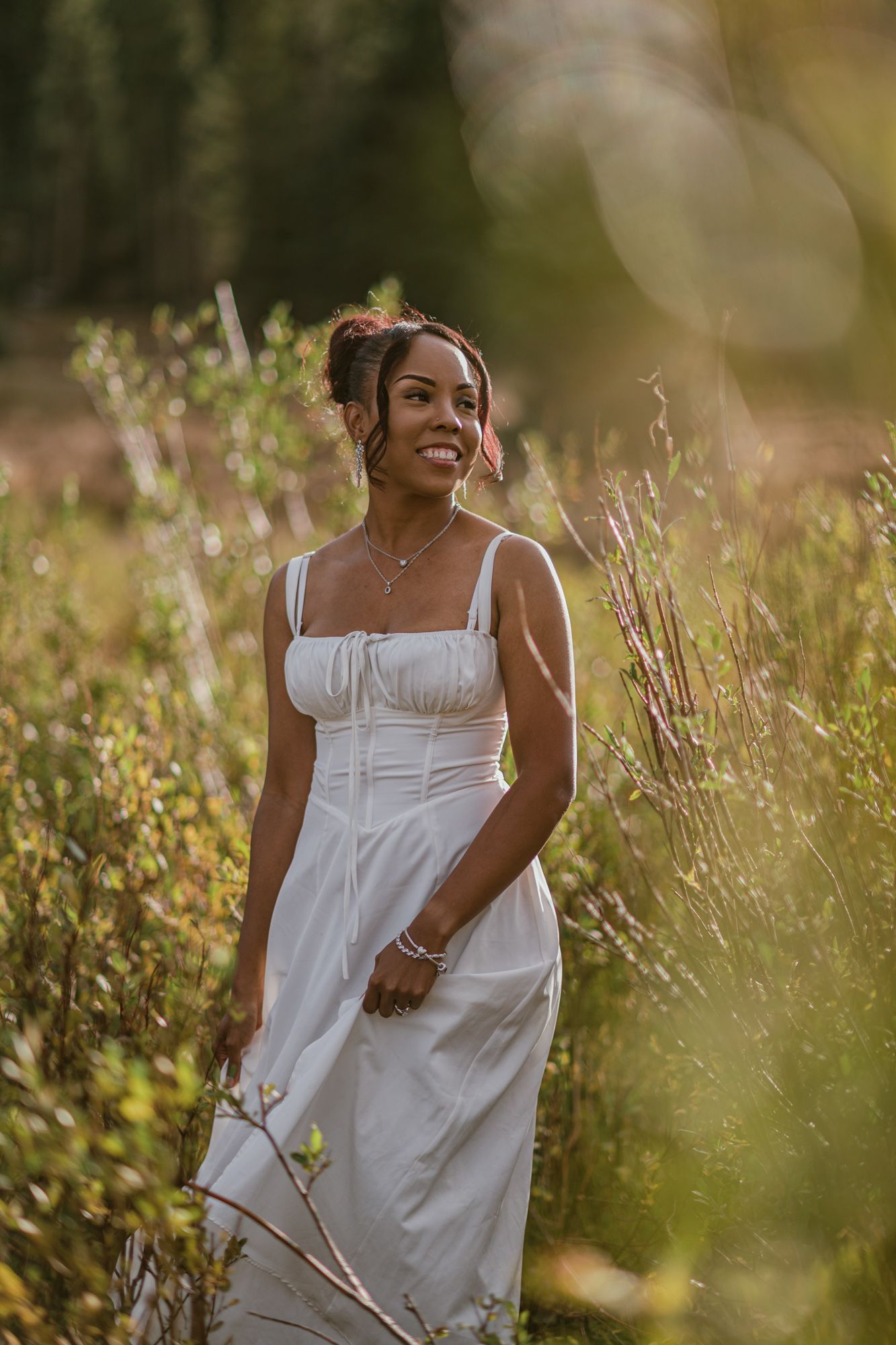 A smiling woman in a white sundress stands among wildflowers in golden sunlight.