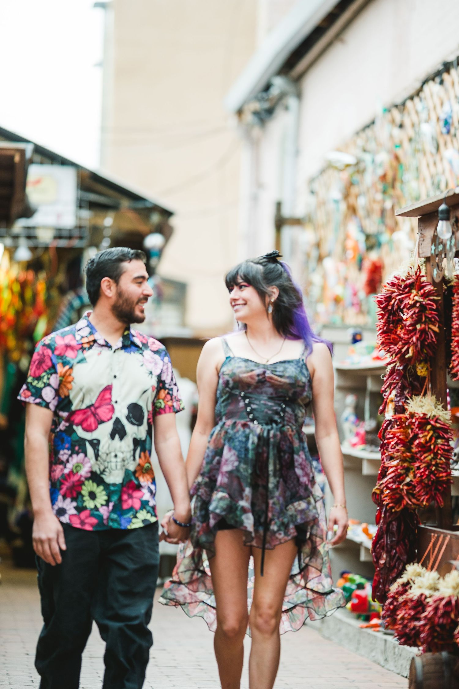 Couple holding hands and smiling while walking through a colorful outdoor market with chili peppers and souvenirs.