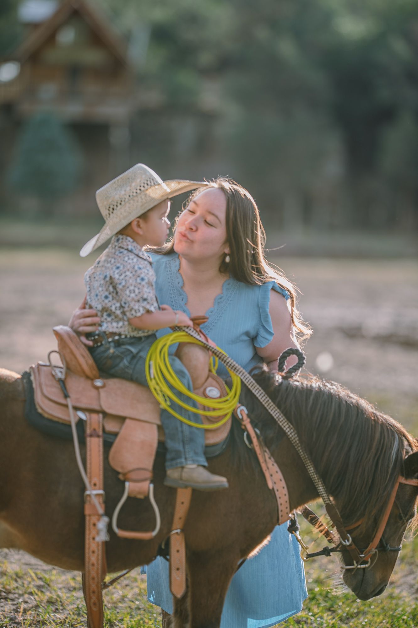 A mother in a blue dress kisses her young cowboy son sitting on a horse, holding a lasso rope outdoors.