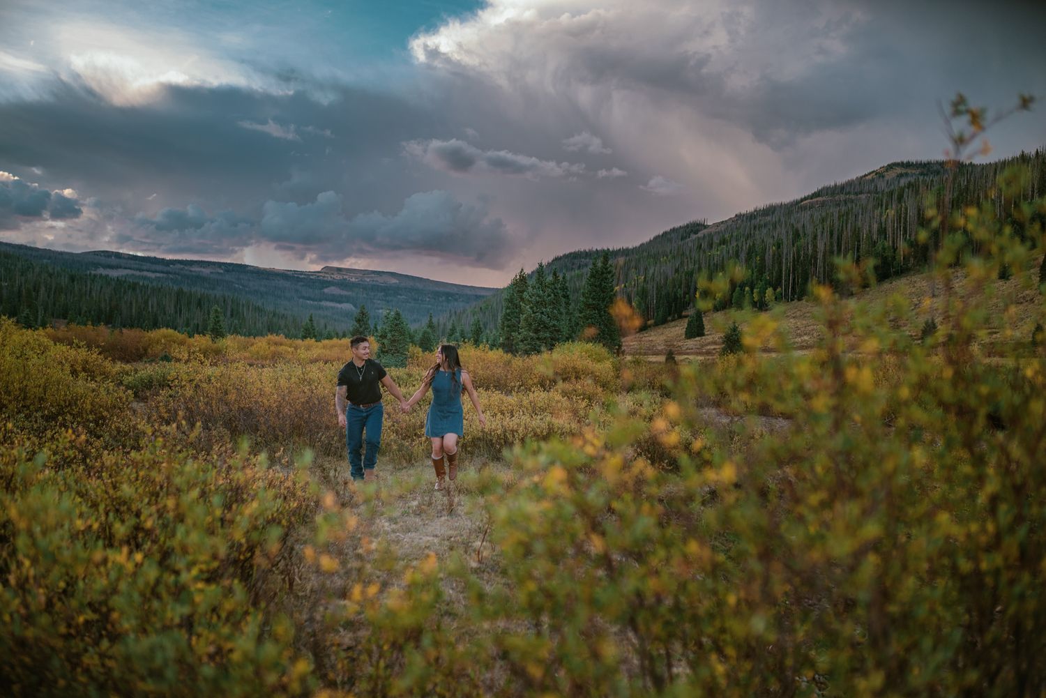 Couple holding hands walking on a mountain trail surrounded by autumn foliage and dramatic cloudy skies.