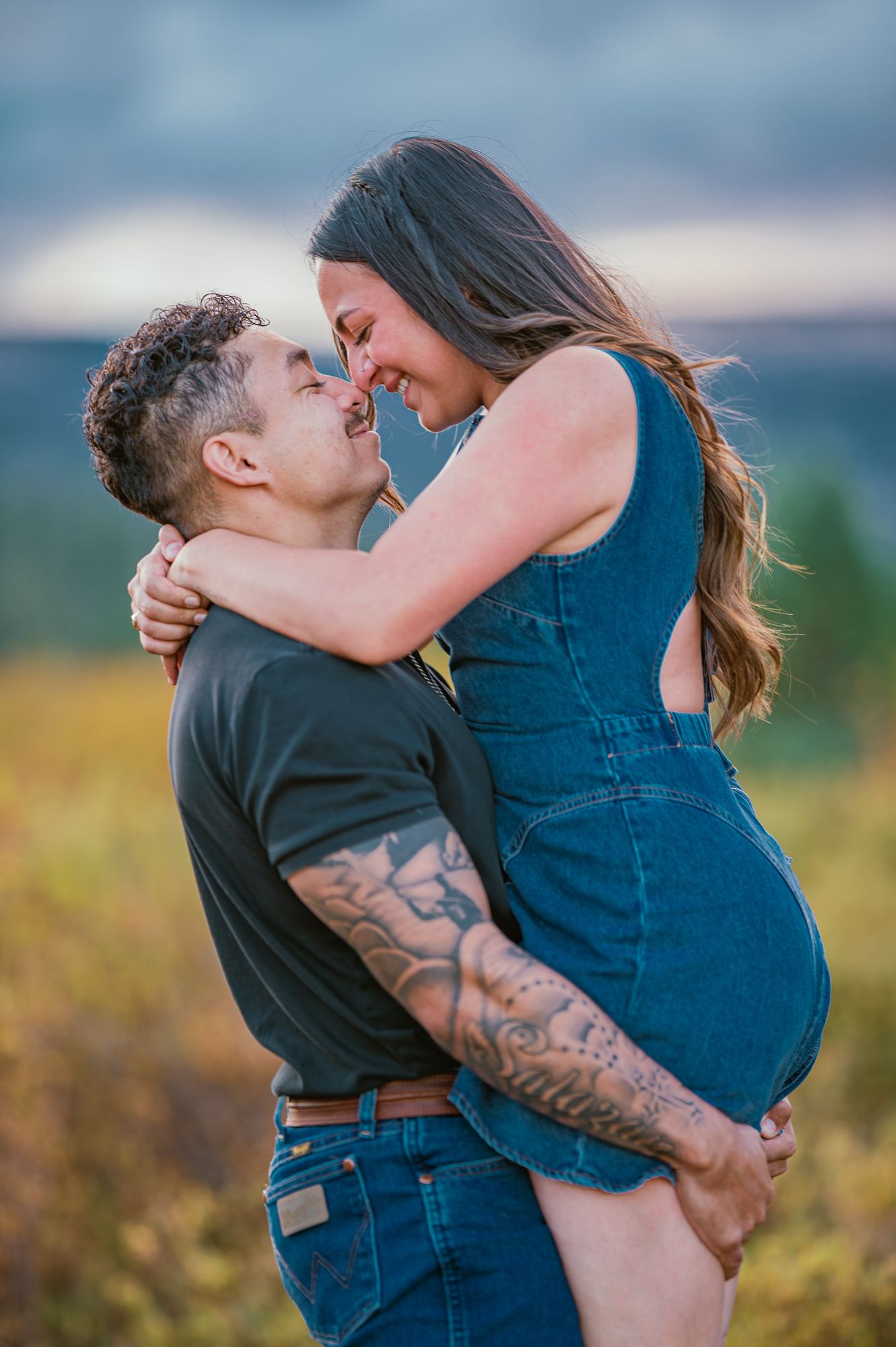 A couple shares a romantic nose-to-nose moment outdoors, man with tattoos lifting woman in denim dress against scenic backdrop.