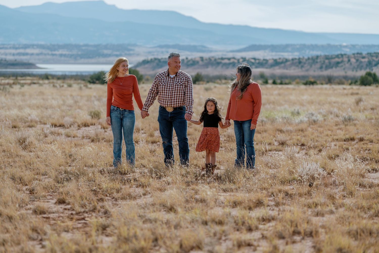 A family holding hands walks together through a golden field with mountains visible in the distance.