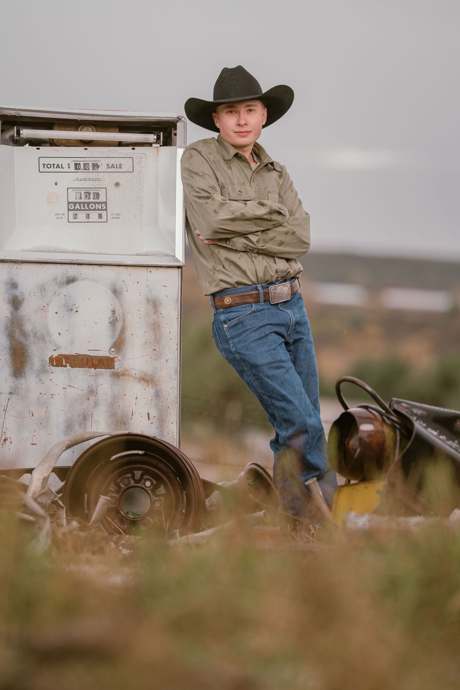 A cowboy in traditional western attire leans against an old white trailer in a rustic outdoor setting.