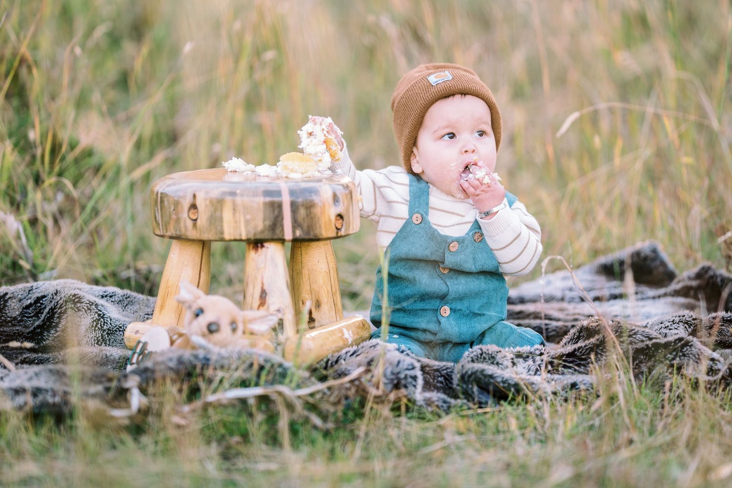 A small child in teal overalls snacks at a rustic wooden table outdoors.
