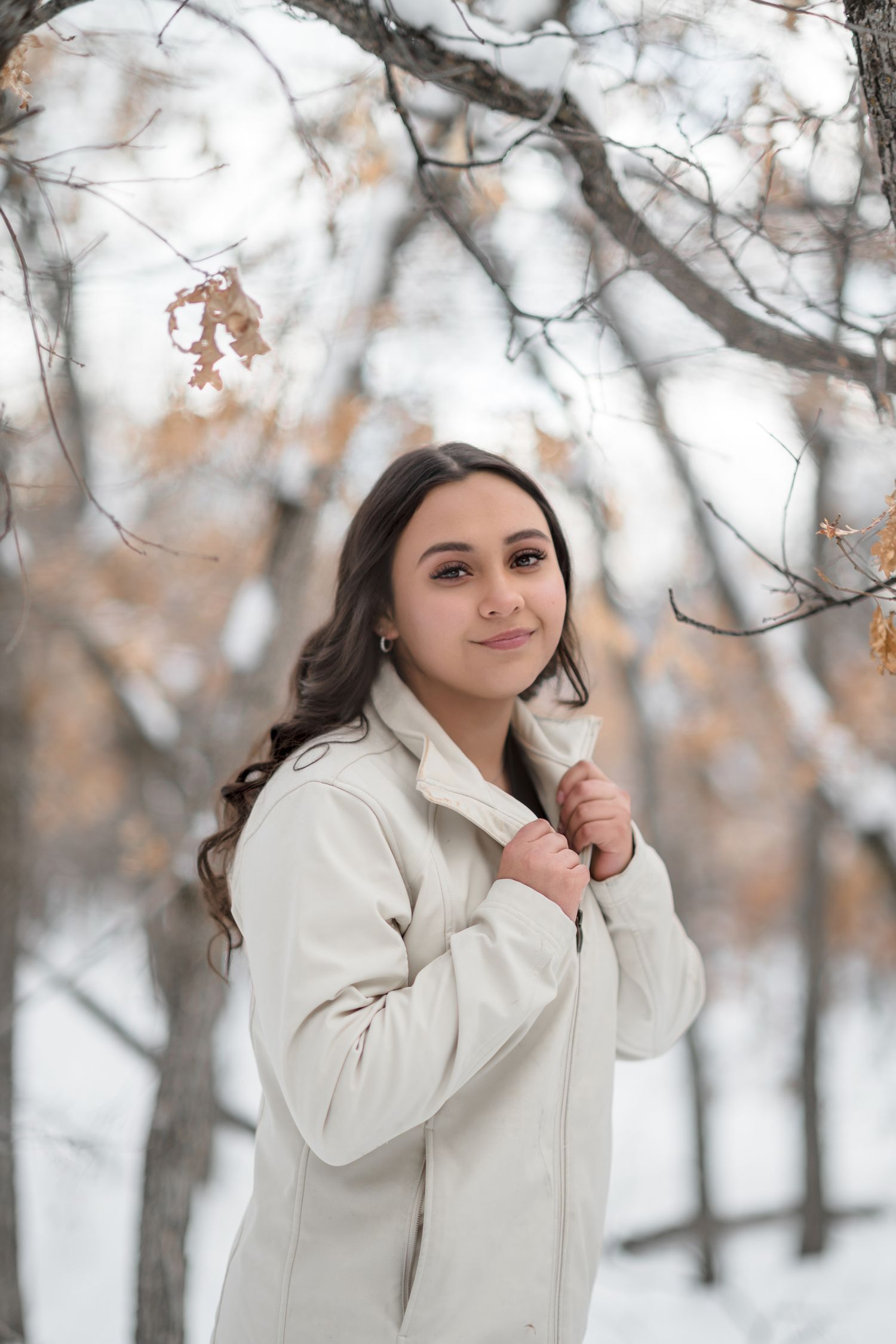 A person in a cozy white winter coat poses playfully in a snowy forest setting with bare trees in the background.