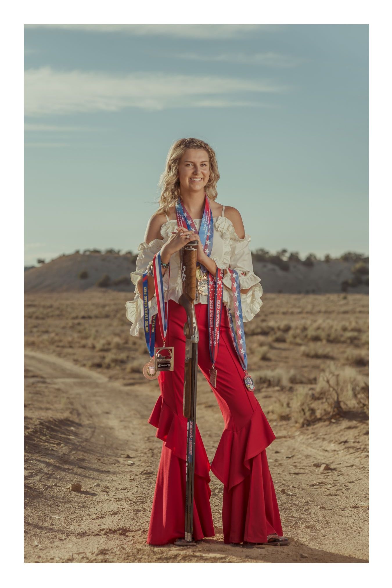 Woman in red flare pants and white blouse holding a guitar on a dusty outback dirt road under blue sky.