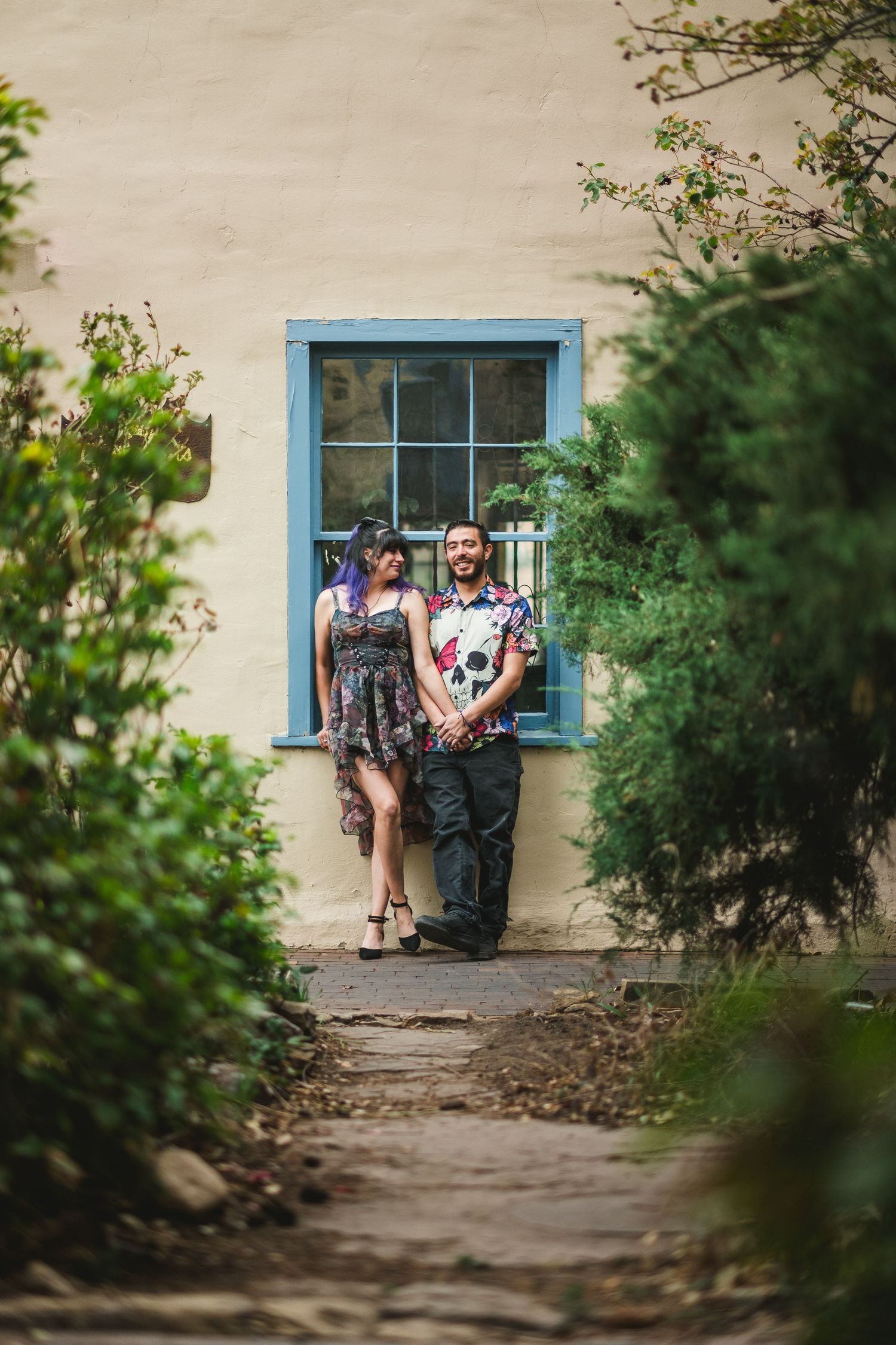 Couple posing together in a garden pathway beside a cream wall with blue-framed window, surrounded by lush greenery.
