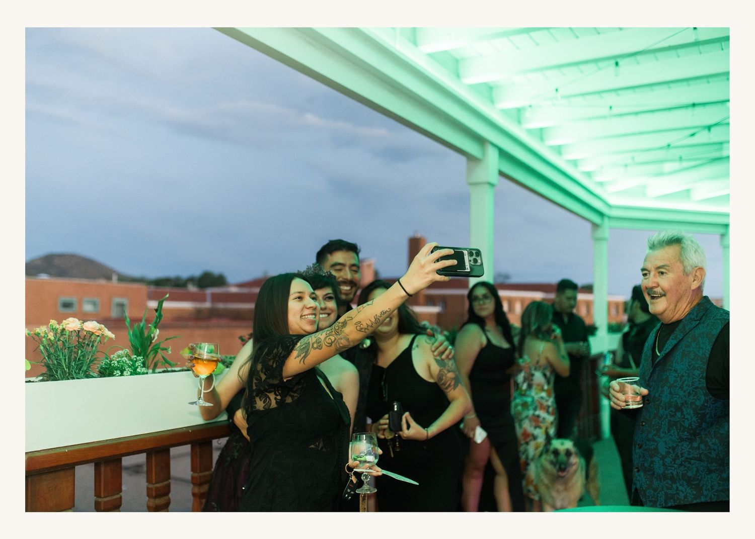 Group of guests taking a selfie at an outdoor rooftop party event under green-lit pergola at dusk.