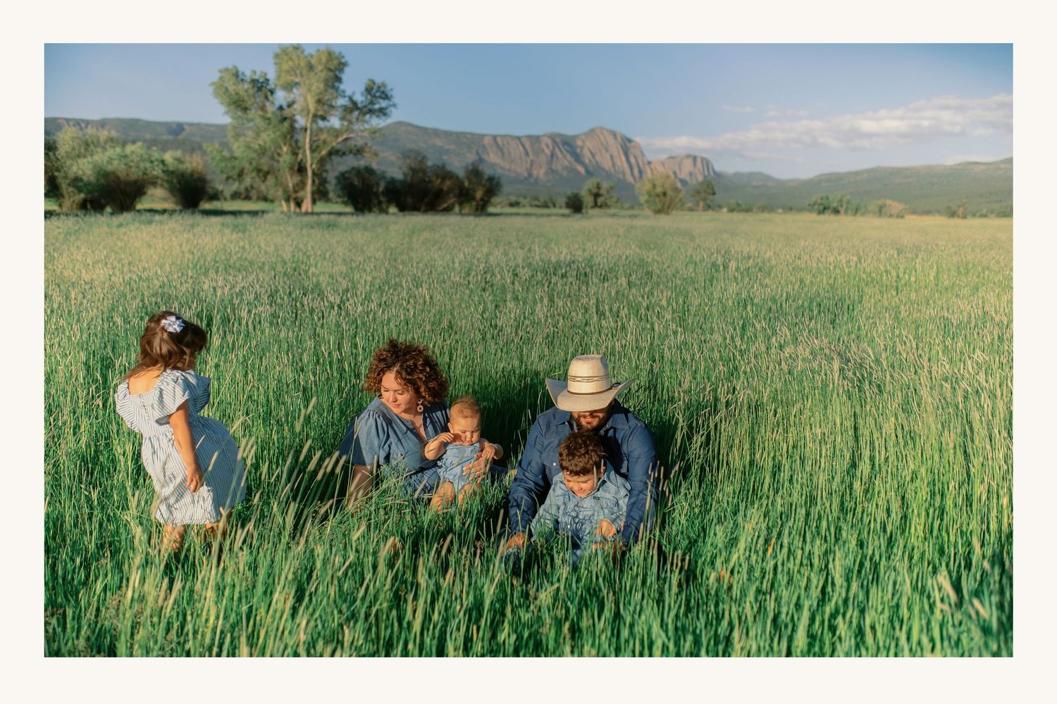 Family with children sitting in tall green grass field with scenic mountain views.