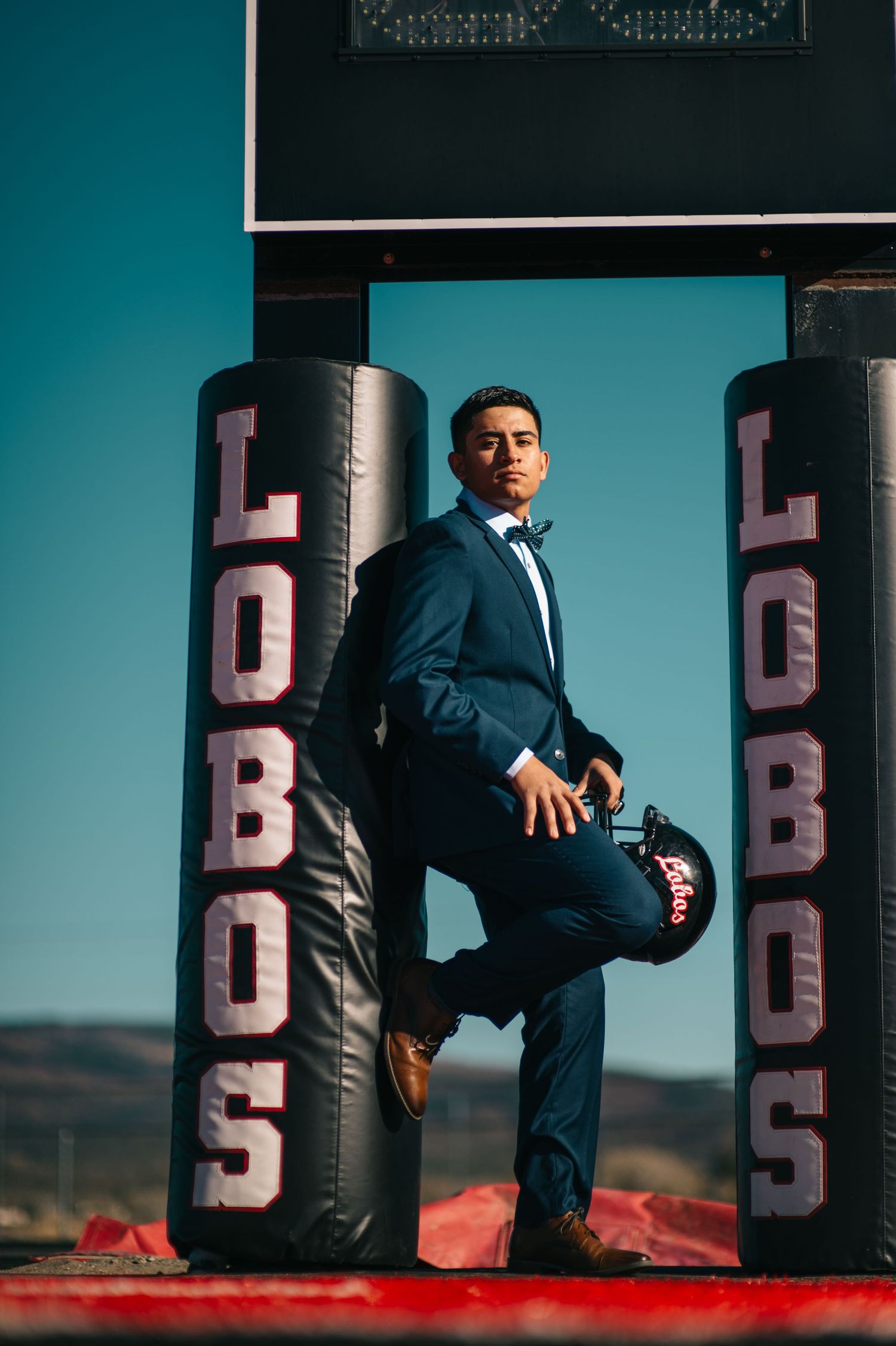 Young man in navy suit posing between Lobos football goal post pads on a sunny day.