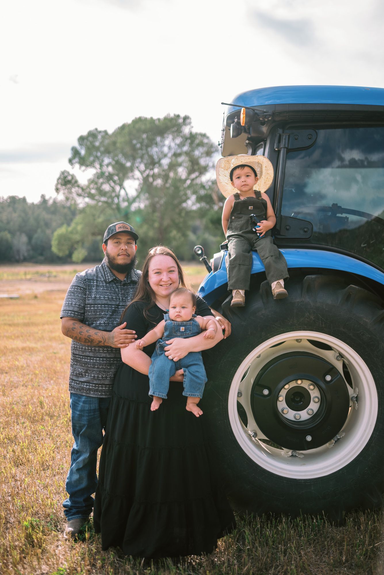 Family of four posing by a blue tractor in a farm field, parents holding baby while toddler sits on tractor tire.