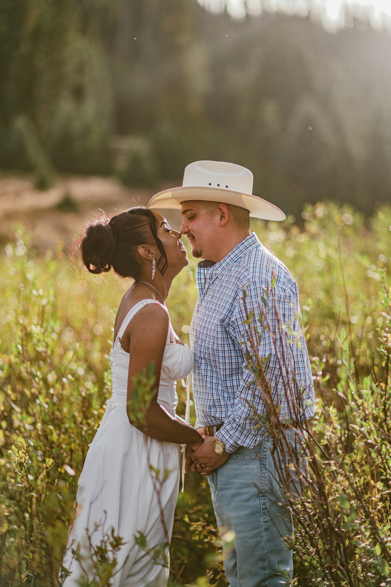 Couple sharing a romantic moment in a sunlit meadow, man in cowboy hat and plaid shirt, woman in white dress.