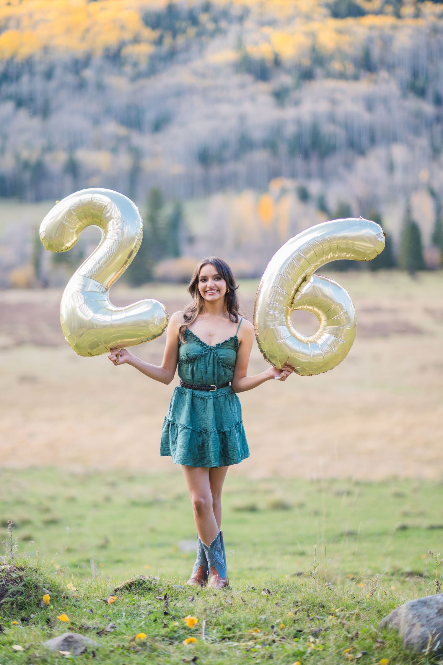 Person in a teal dress holds gold number balloons while standing in a scenic mountain meadow with autumn colors.