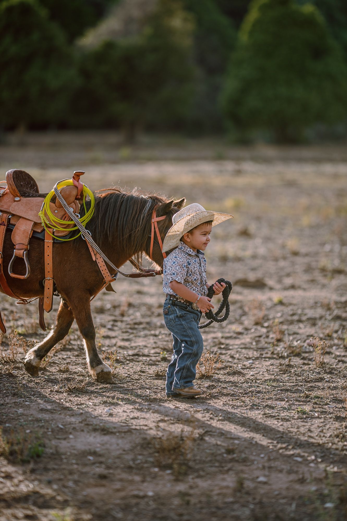 Young child in cowboy hat and jeans leading a saddled pony across a dusty field at golden hour.