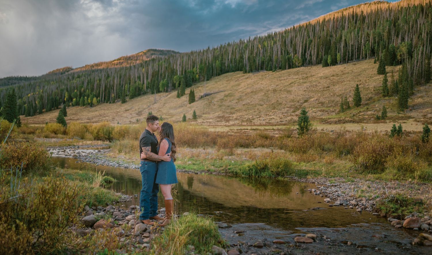 Couple sharing a romantic kiss by a mountain stream in a scenic Colorado valley with pine trees and golden meadows.