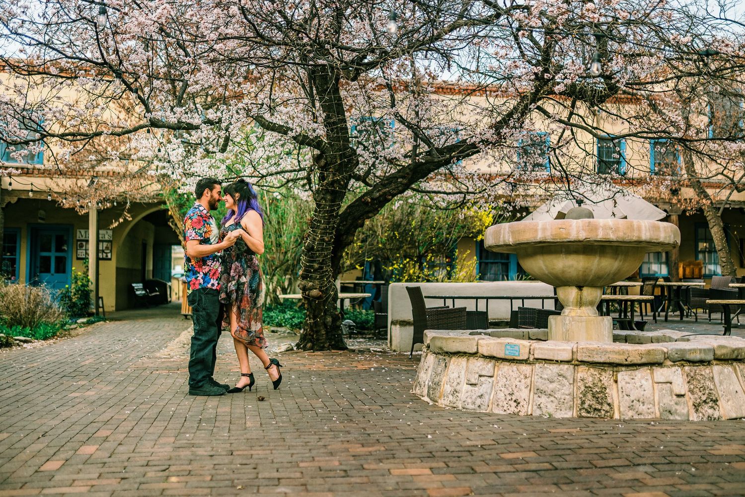 Couple kissing near a stone fountain in a brick courtyard with blooming cherry blossom trees in spring.