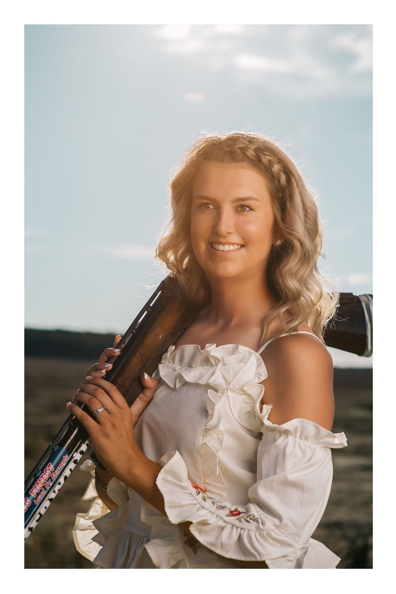 Smiling young woman in white off-shoulder top holding a shotgun outdoors under a bright sky.