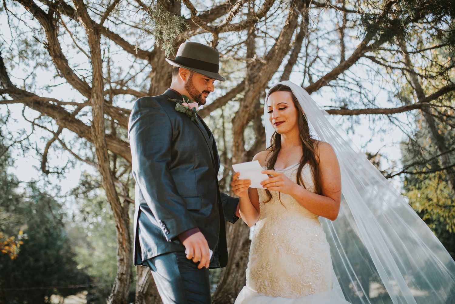 Couple reads wedding vows under sunlit trees during outdoor ceremony.