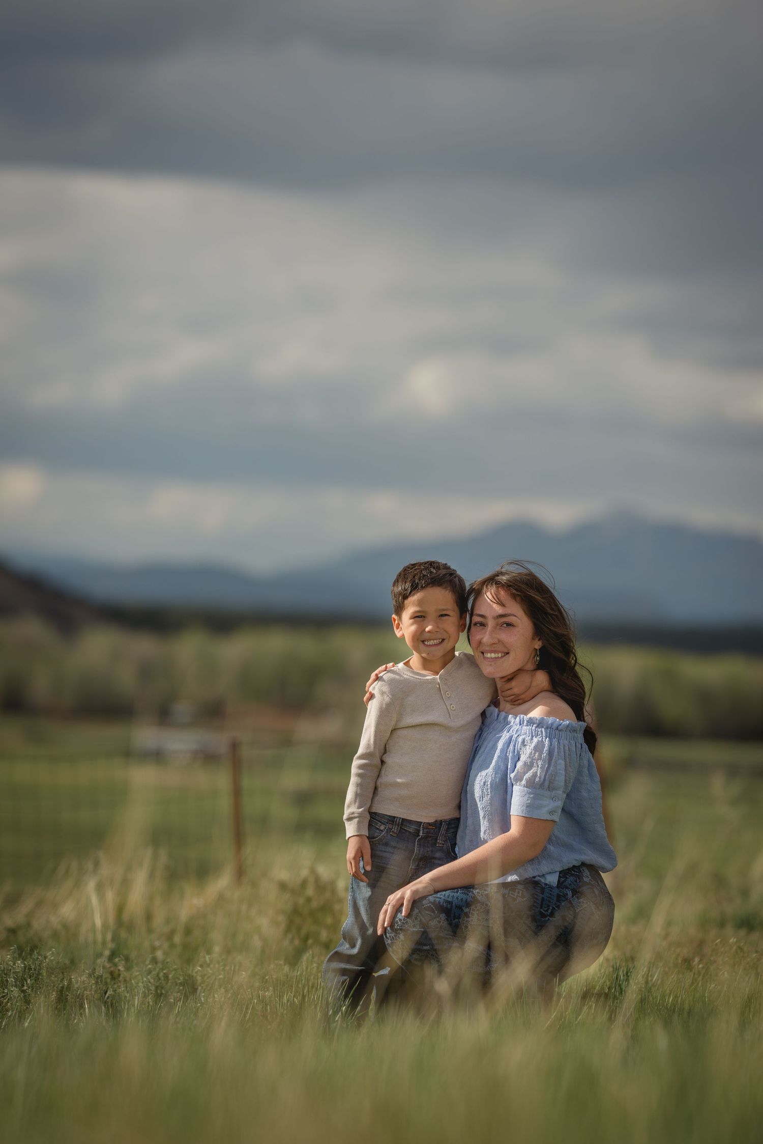 Two people share a tender moment together in a grassy meadow with mountains and dramatic cloudy sky in the background.