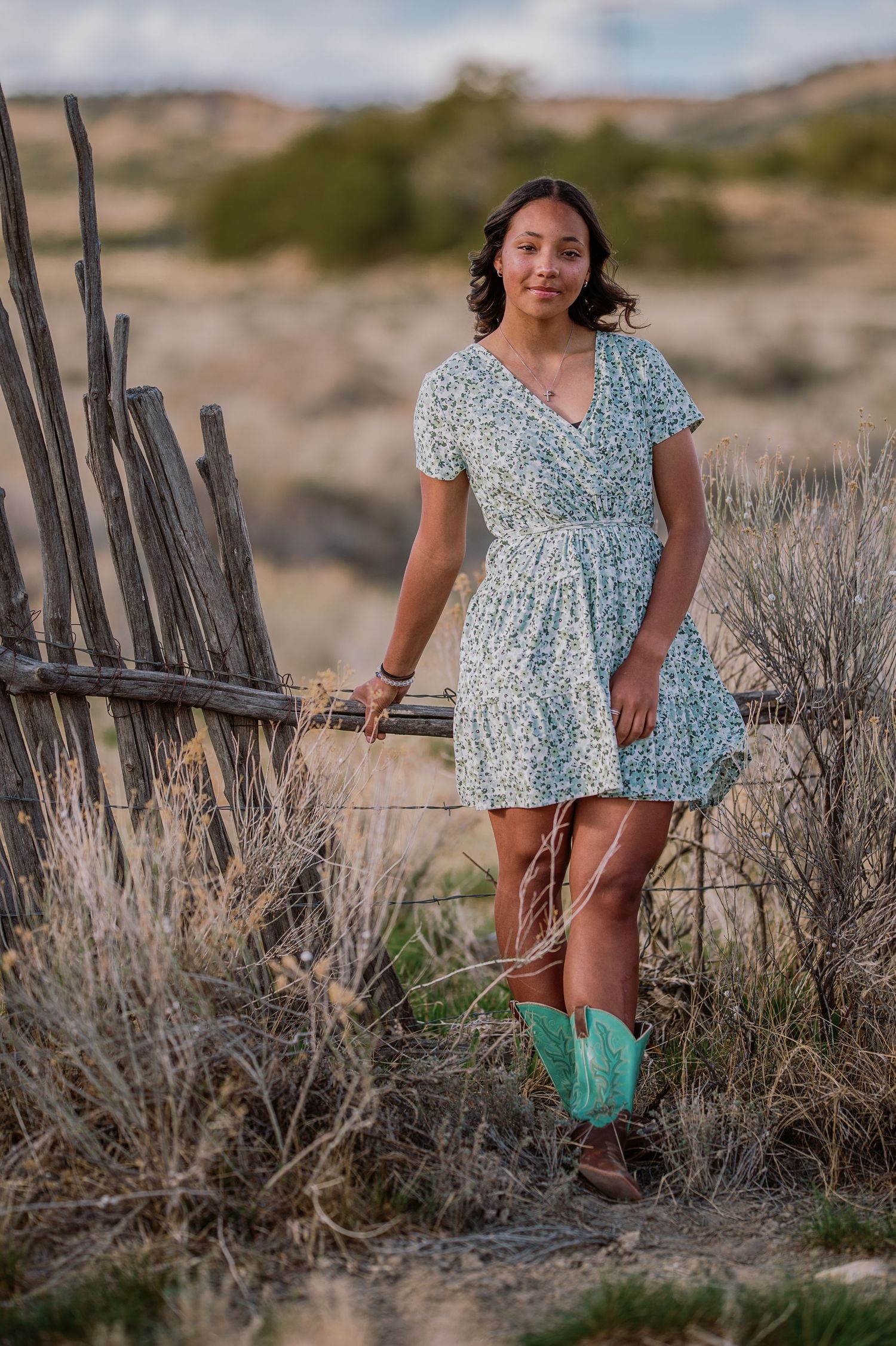 A person in a floral dress and green boots stands in desert brush near wooden fence posts at sunset.
